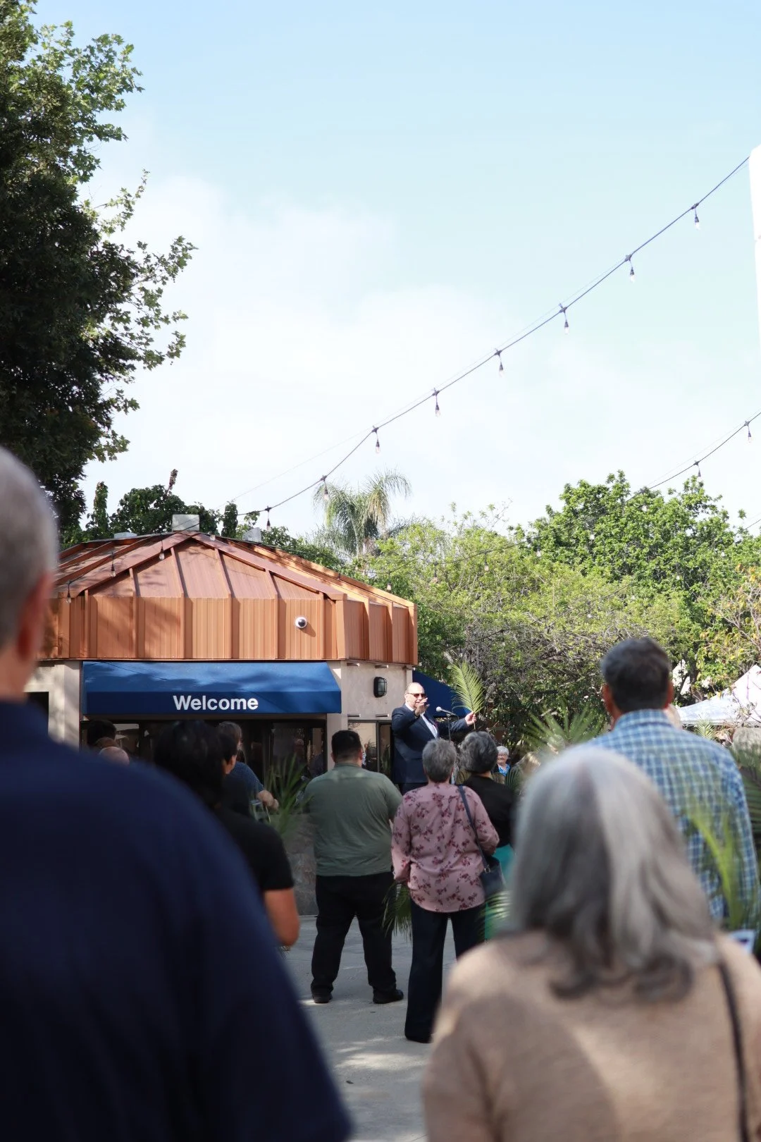 A group of people gathered outdoors listening to a speaker in front of a small building with a blue 'Welcome' sign. The speaker is dressed in formal attire and is holding a microphone, gesturing with his other hand. Trees and string lights are visible overhead.