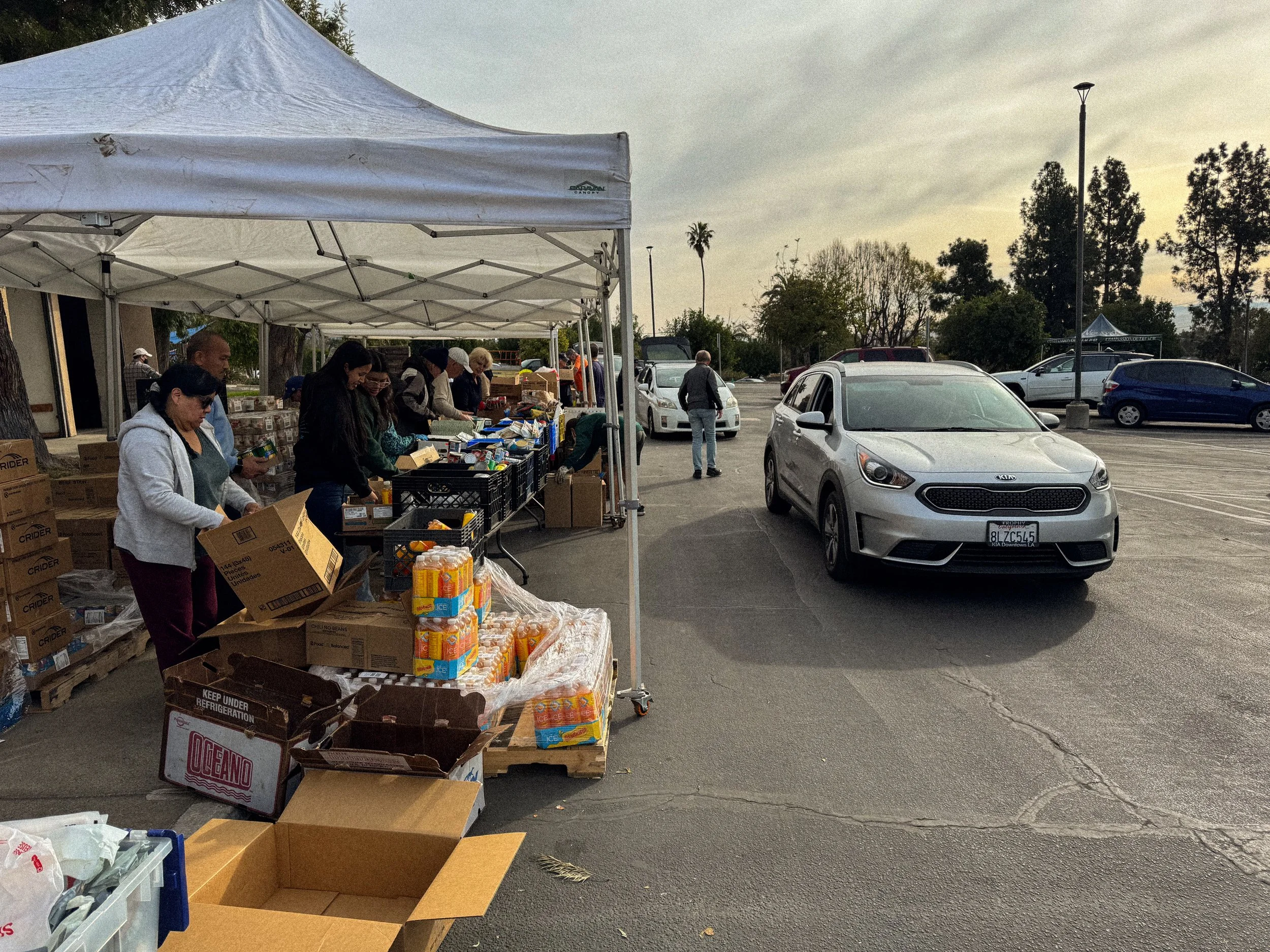 People arranging and selling items at a daytime outdoor market stall with a white canopy, in a parking lot with parked cars and trees in the background.
