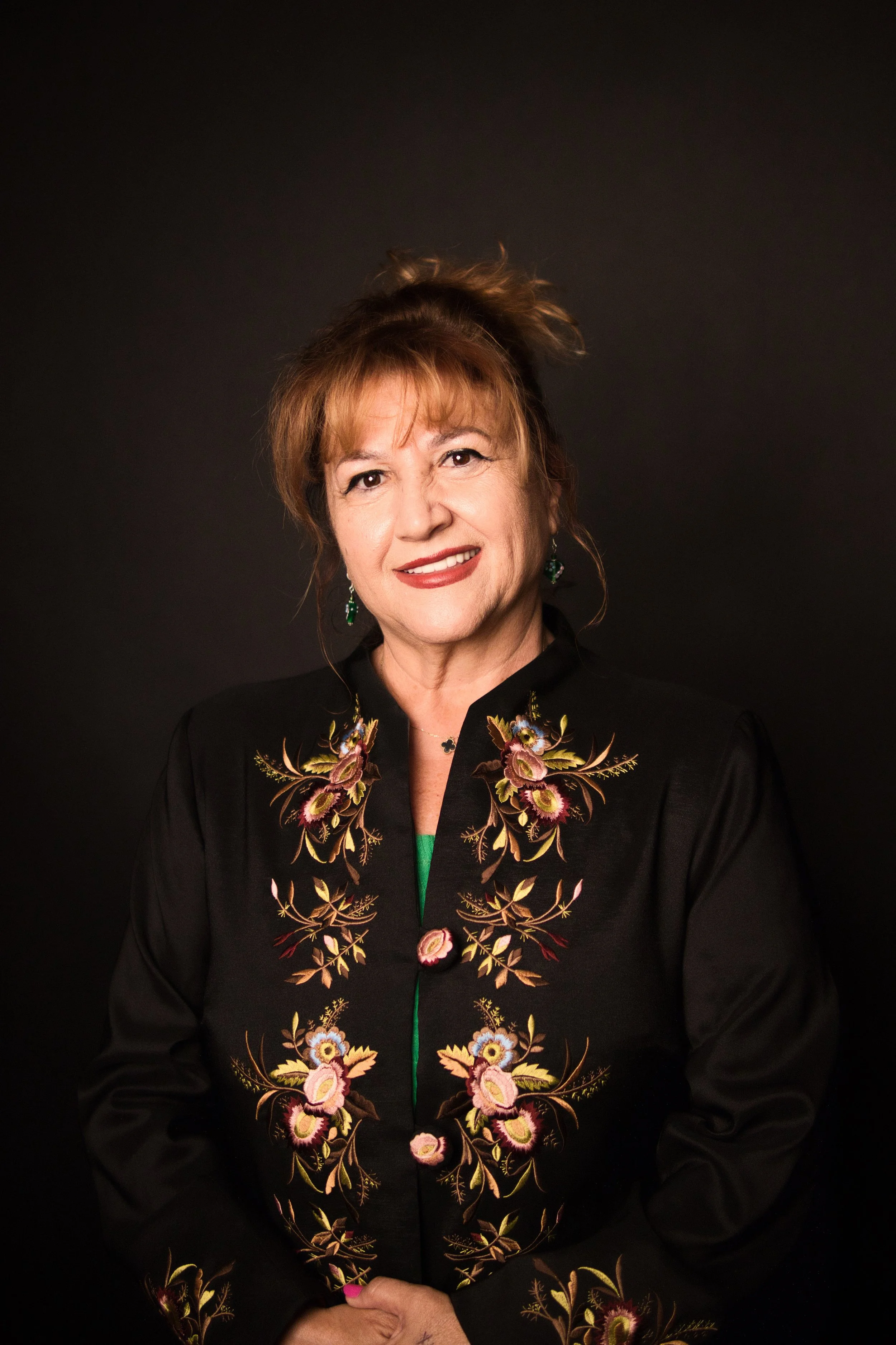 A woman with red hair in a top knot bun, wearing earrings and a black embroidered jacket with floral and animal designs, smiling slightly against a plain dark background.