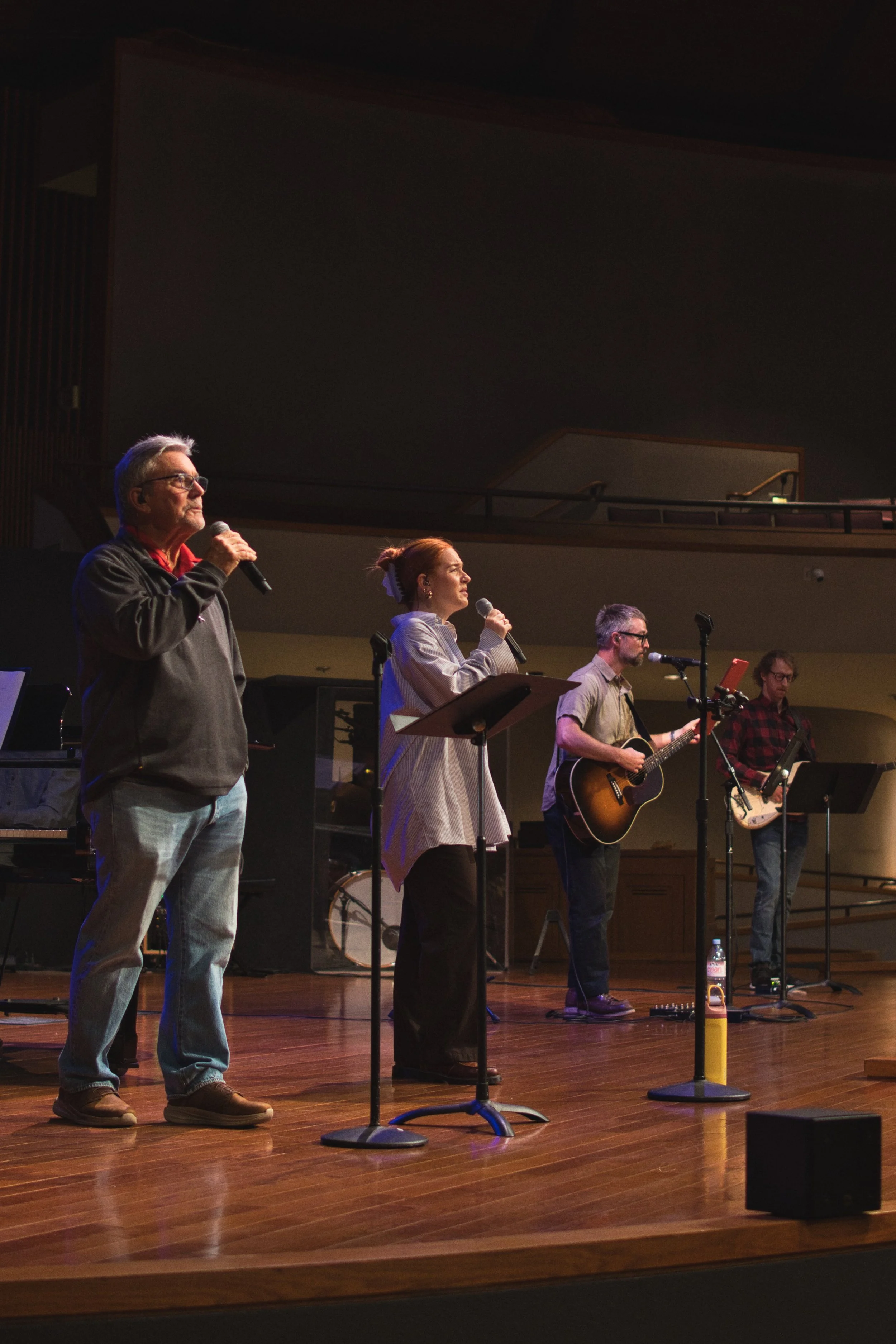 A group of five people singing worship music on a stage at church. One person is playing guitar, and there are music stands and equipment around them, in a large room with a high ceiling and balcony.