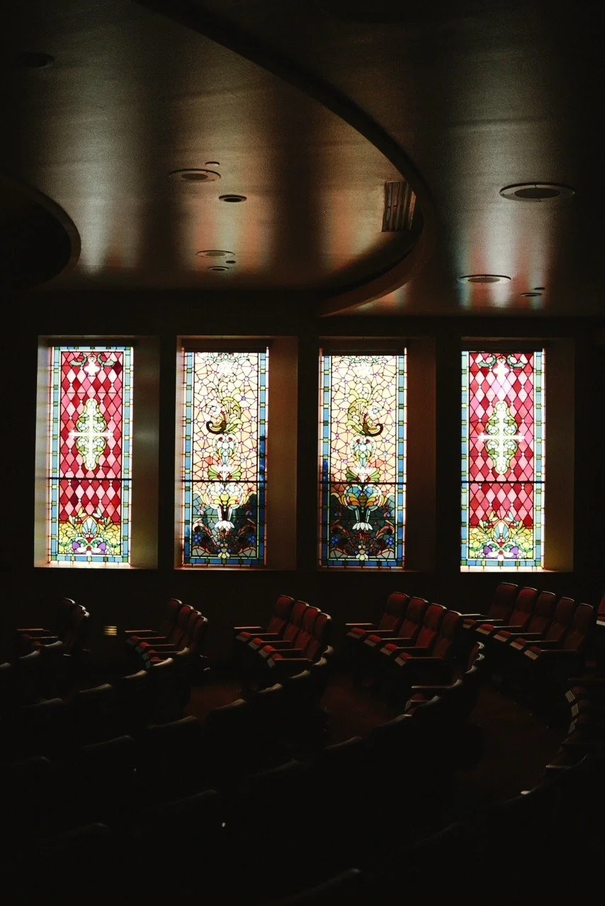 Inside a dimly lit room with four stained glass windows and rows of empty red cushioned chairs.