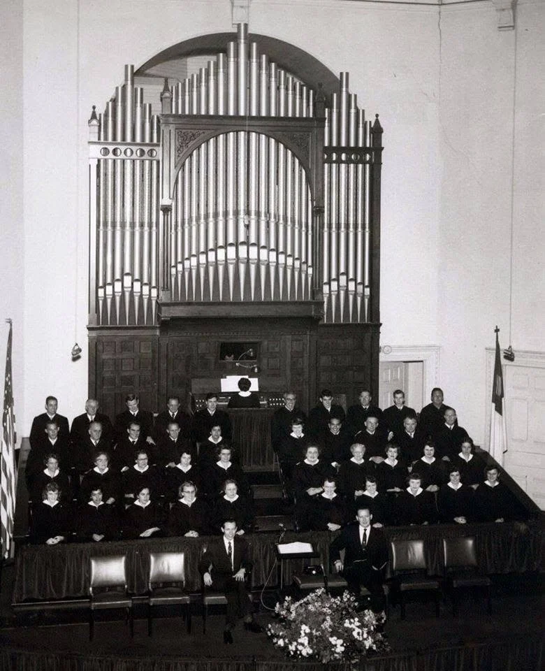 A choir of people in robes standing on risers in front of a large pipe organ inside a church or auditorium.