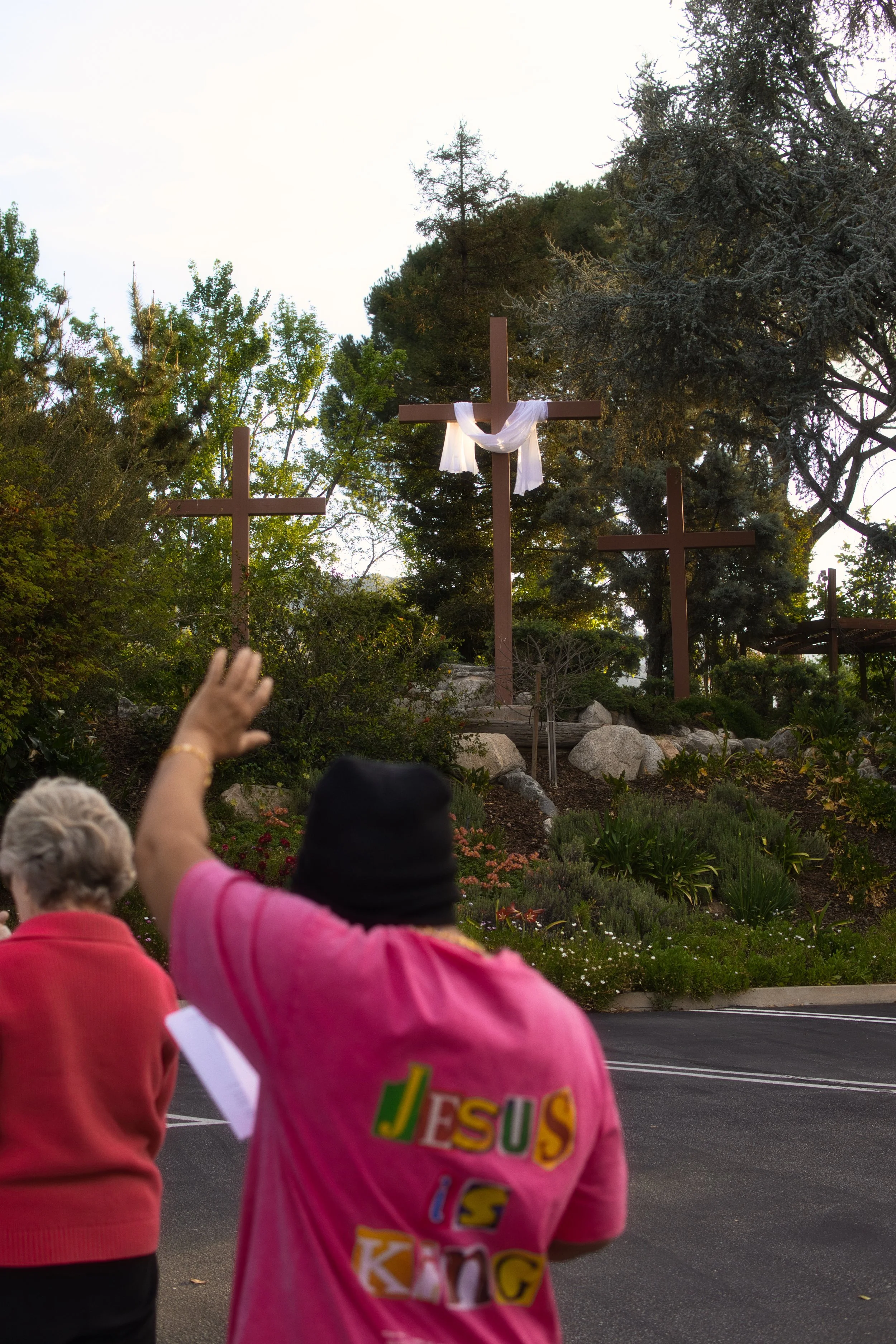 People standing outdoors near religious crosses, with some raising their hands, surrounded by trees and plants.