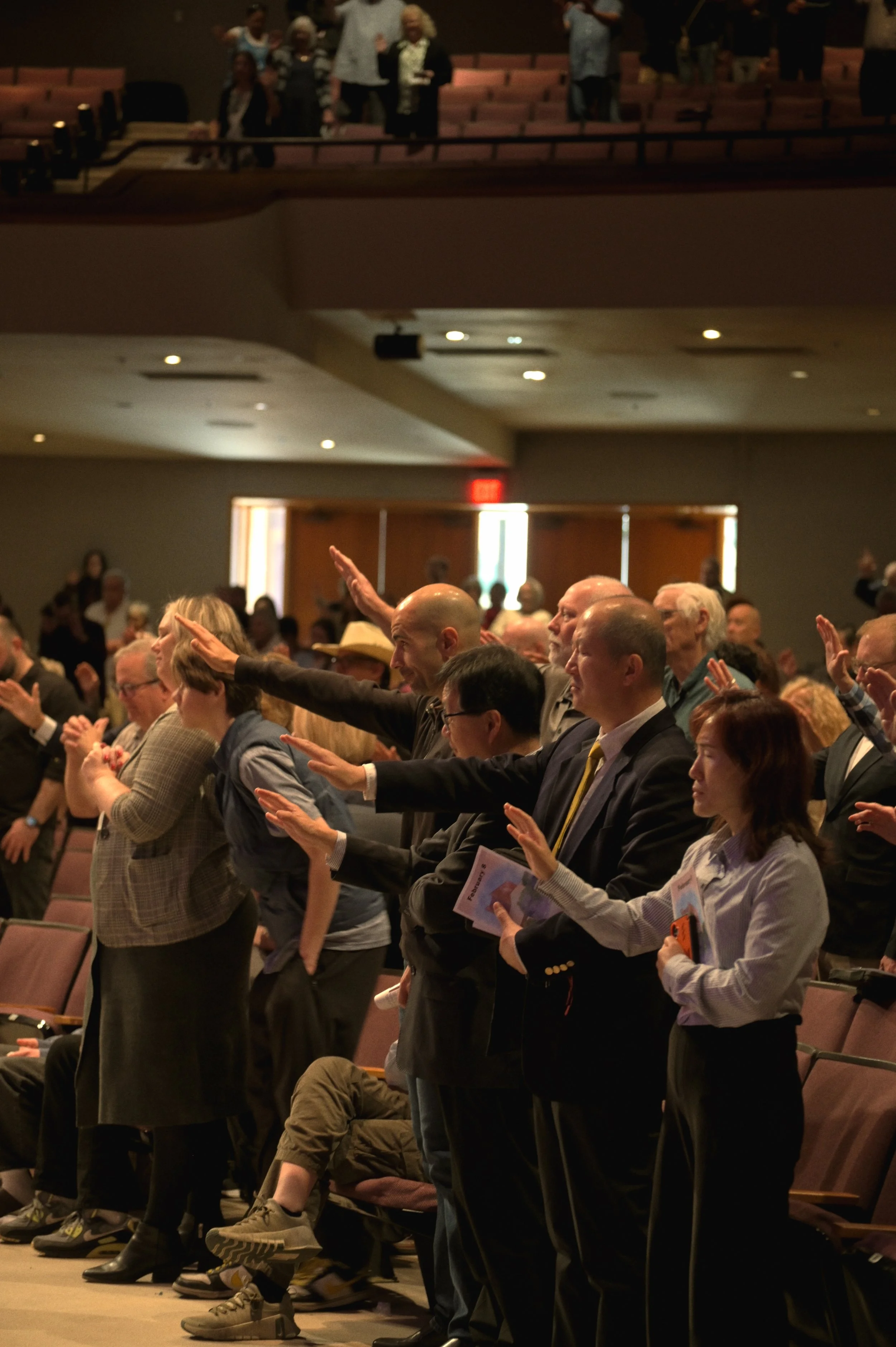 A group of diverse people standing and raising their right hands during a ceremony or pledge in a large auditorium with pink seats and a wooden door in the background.
