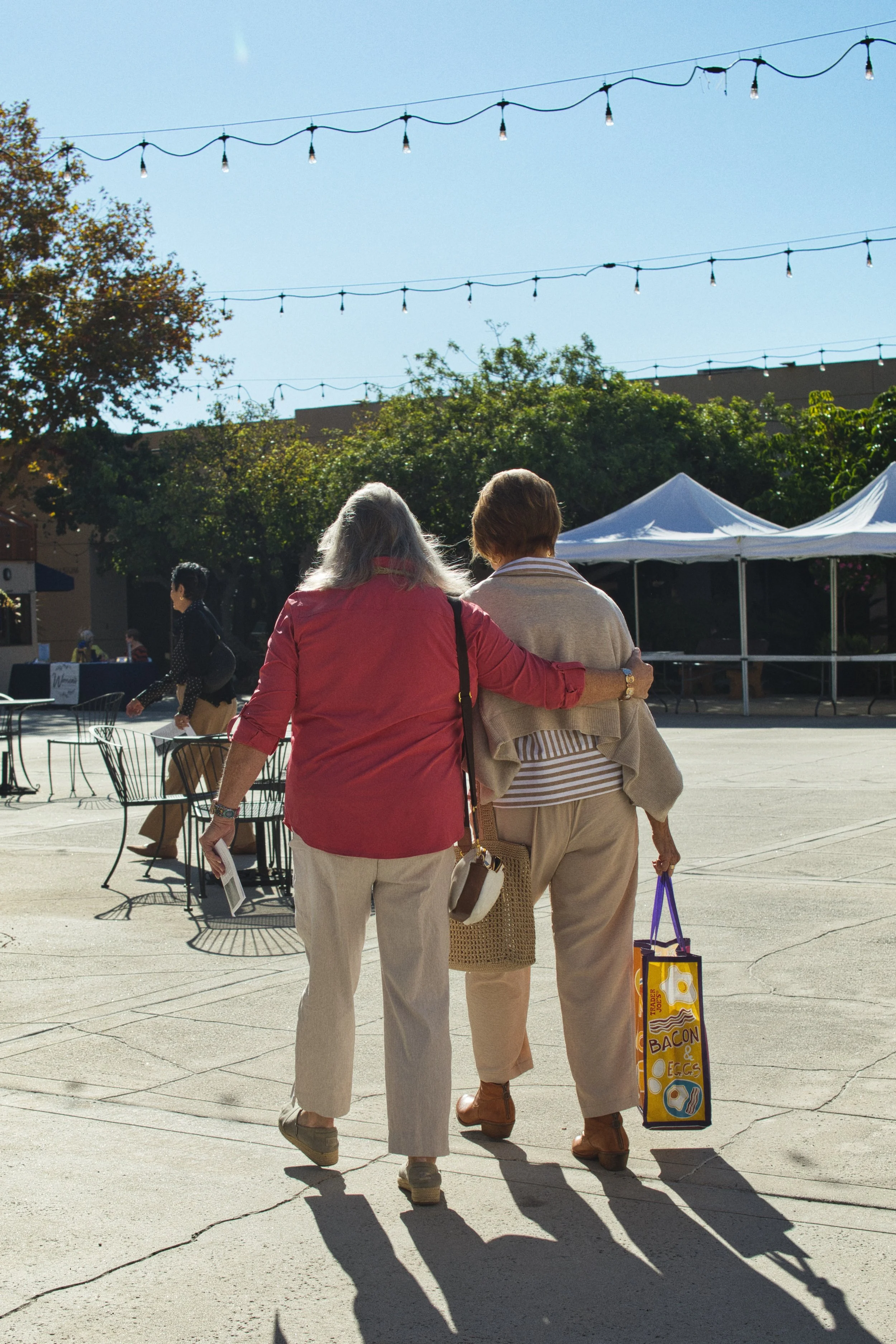 Two women walking arm in arm at an outdoor event, with a market tent and string lights in the background.
