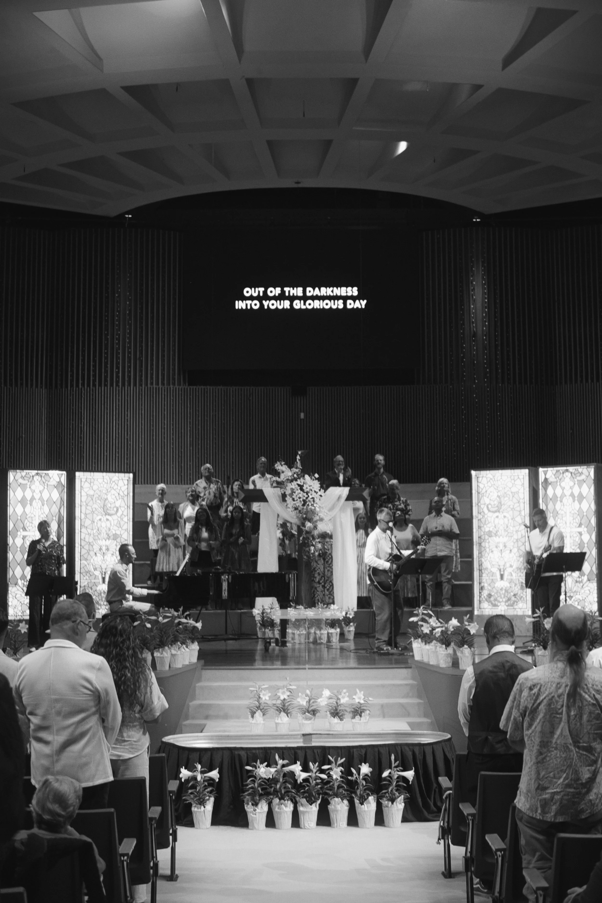 People gathered in a church for a service or event, with a band performing on stage and a large screen displaying the words "Out of the darkness into your glorious day." Potted plants with flowers line the front of the stage.