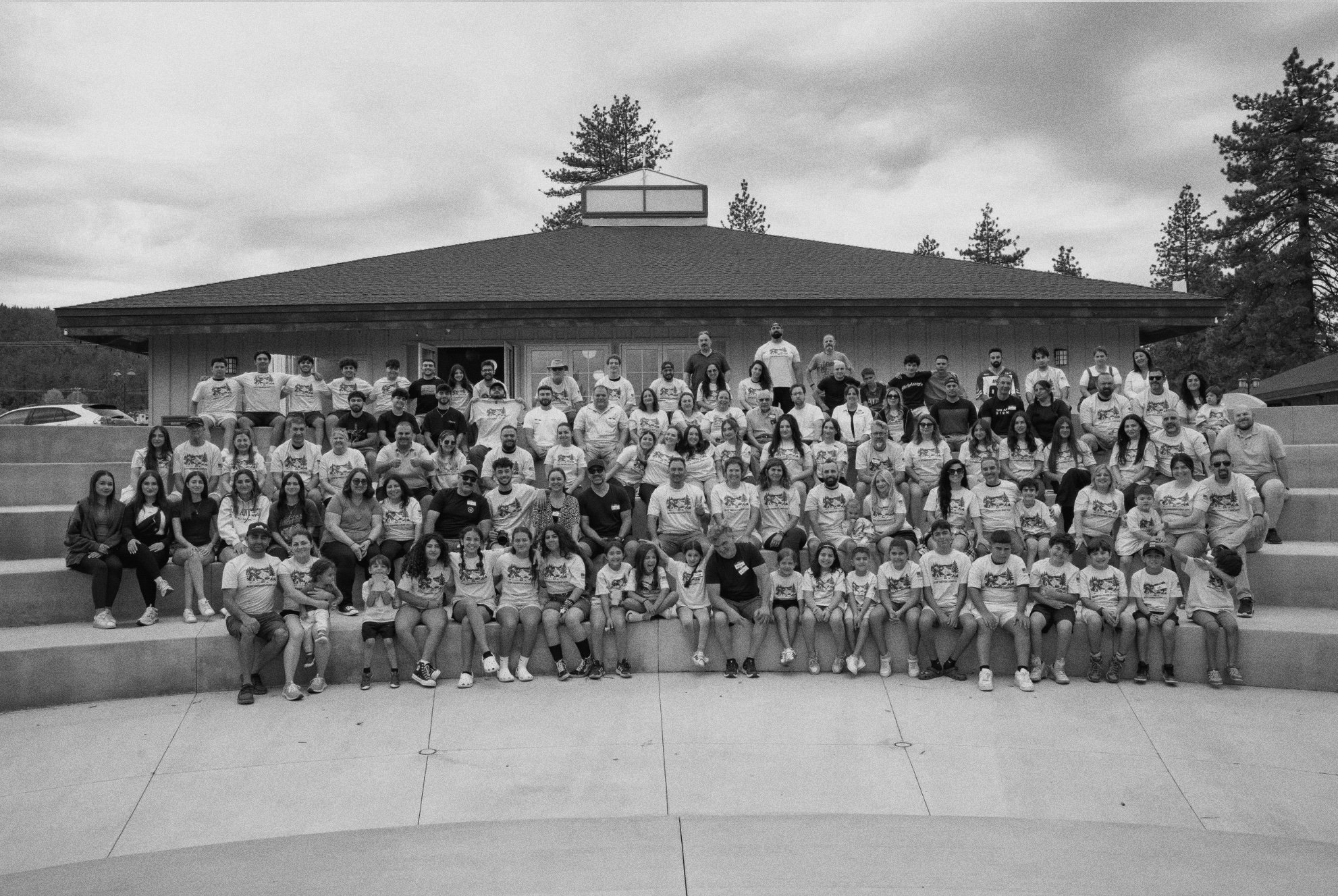 A large group of people, including children and adults, gathered together on the steps in front of a building with a peaked roof and trees in the background. Many are wearing matching T-shirts with a logo or graphic.