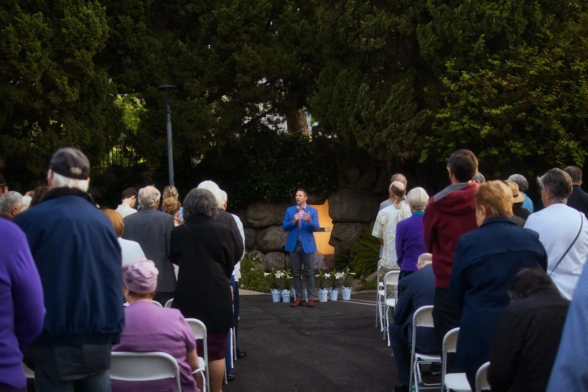 A Pastor in a blue blazer giving a sermon at the church's sunrise Easter service with an audience of diverse people sitting on white chairs, surrounded by lush green trees.