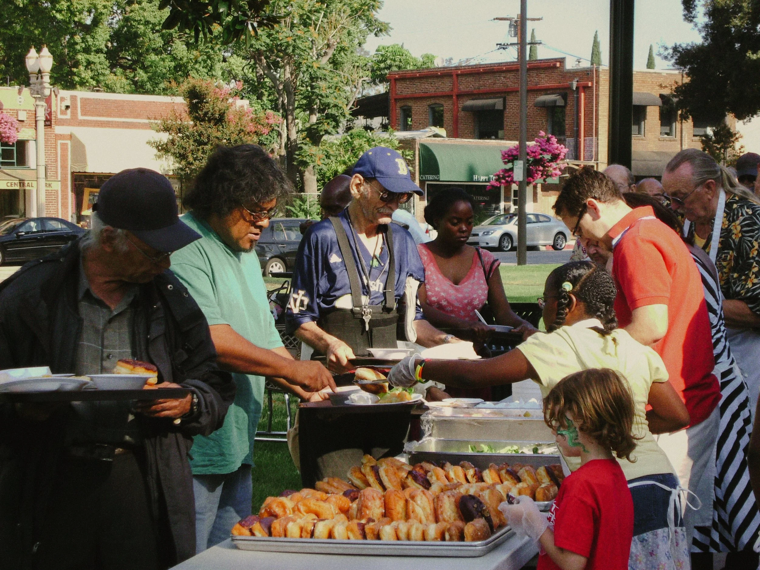 People gathered around a table with trays of food at an outdoor event in a park, with buildings, trees, and a green canopy in the background.