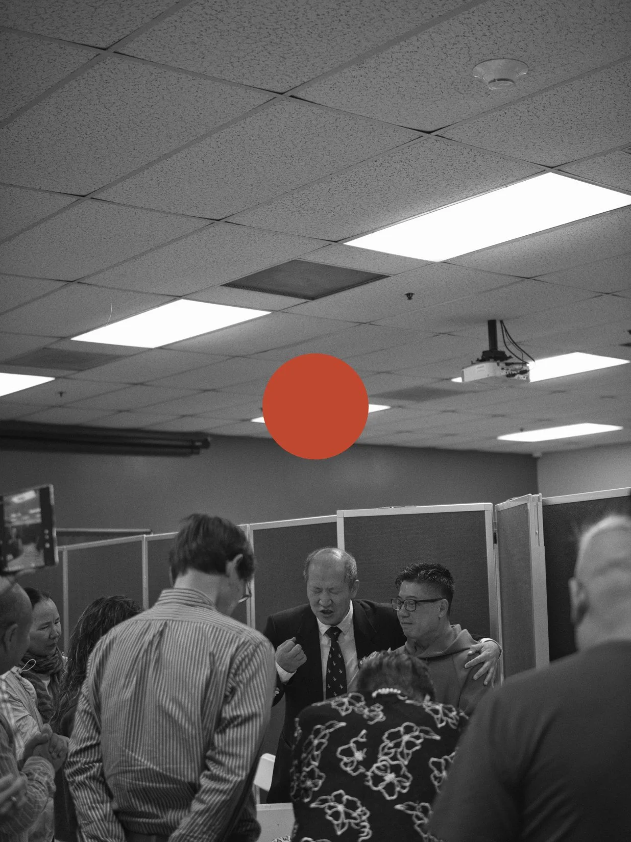 A group of people standing together in an office or conference room, engaged in discussion while a man in a suit appears to express an emotion, with ceiling lights and a ceiling-mounted projector visible in the background.