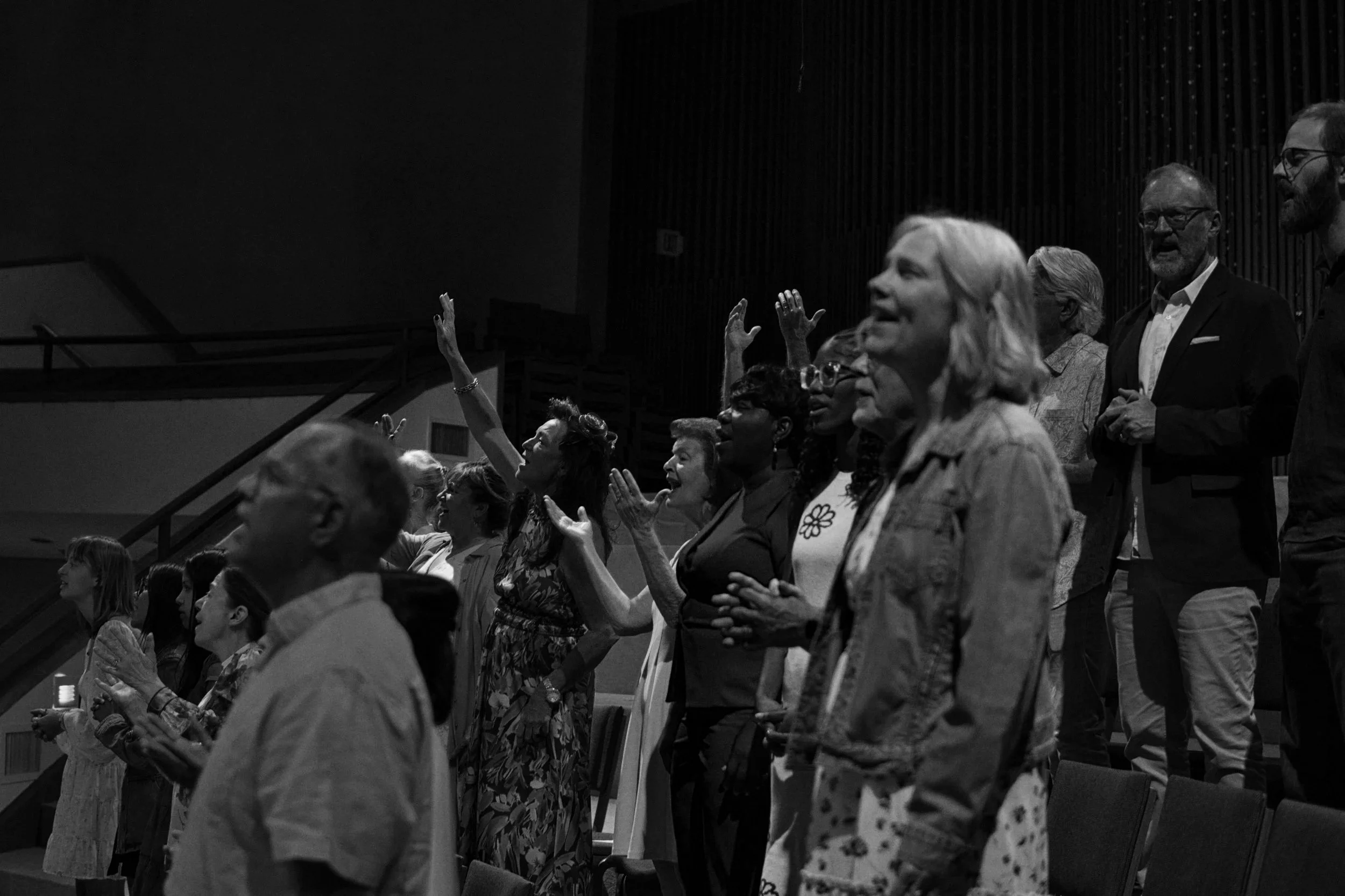 A diverse group of people standing with hands raised in what appears to be a religious or worship service, inside a church or auditorium.