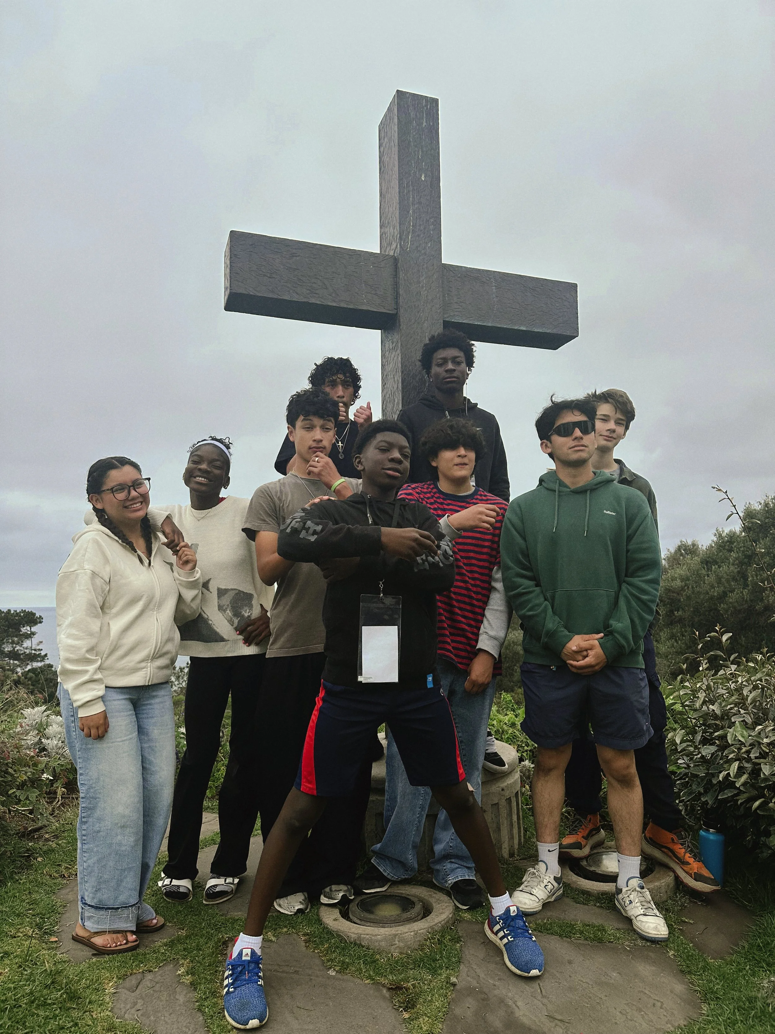 A group of diverse teenagers posing in front of a large cross monument outdoors. The sky is overcast, and the surroundings include green plants and rocks.