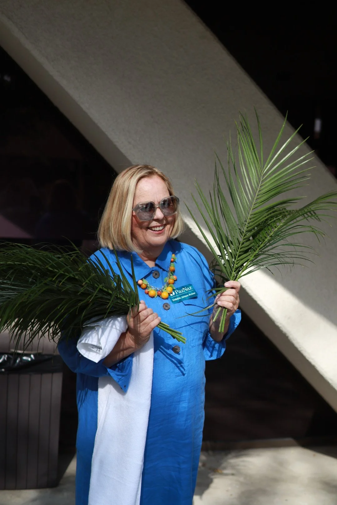 smiling woman with blonde hair, wearing sunglasses, a blue jacket, and a colorful necklace, holding palm leaves.