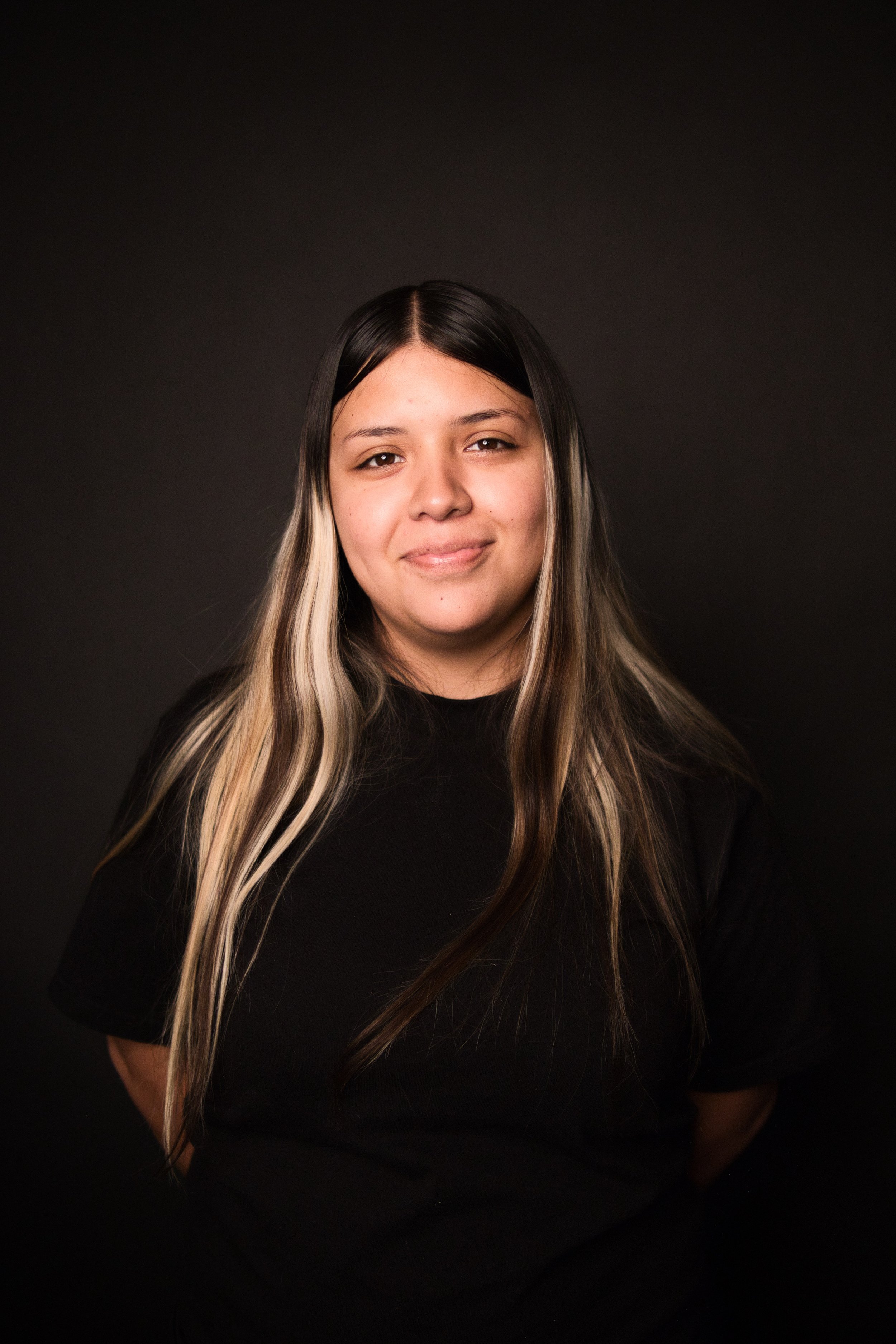 A young woman with long hair, partially blonde and partially black, smiling and wearing a black top, standing against a dark background.