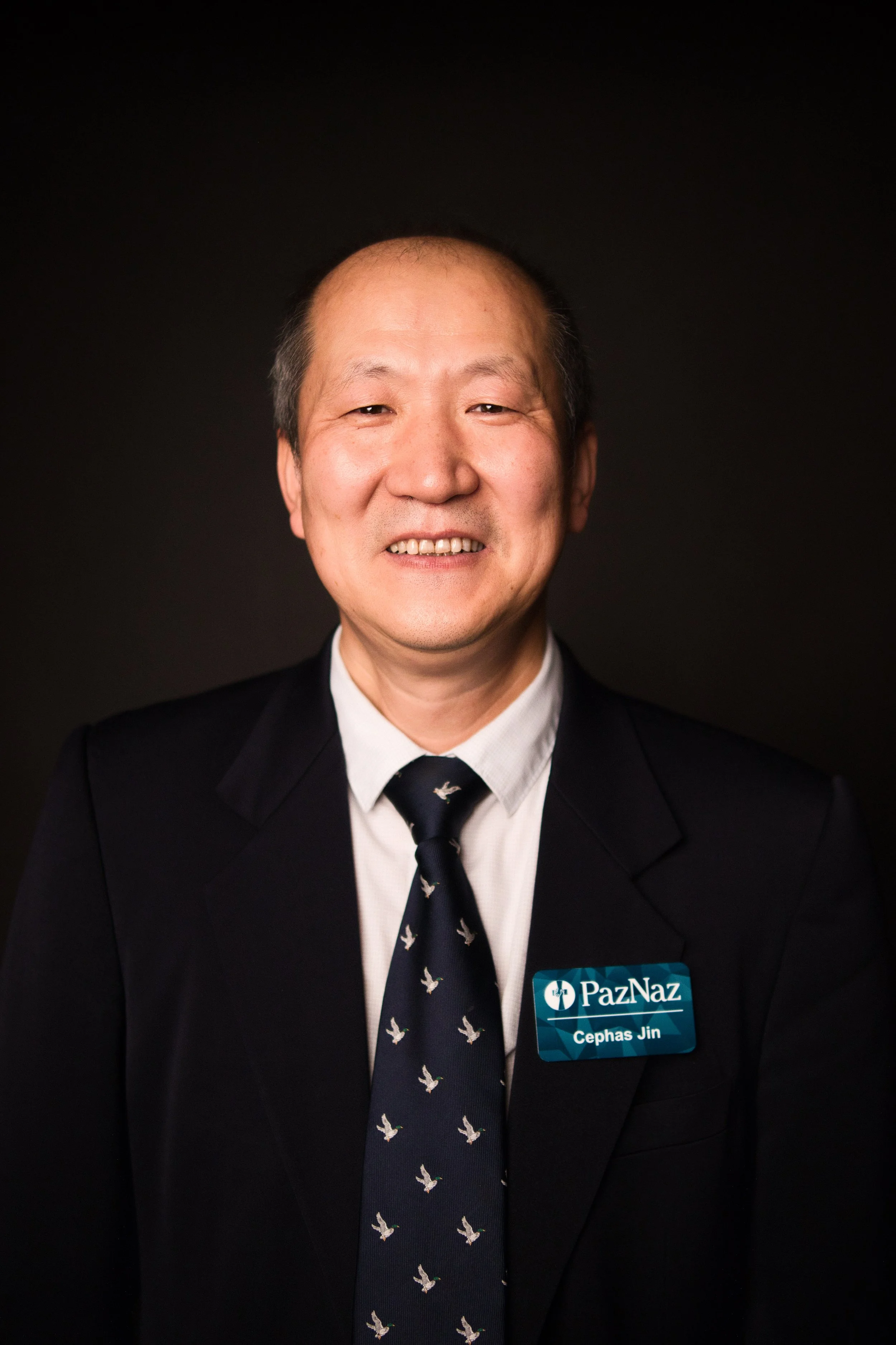 Headshot of a man wearing a black suit, white shirt, and a tie with a bird pattern. He has a name tag that says "Cephas Jin" and a logo with the words "PazNaz". He is smiling against a dark background.