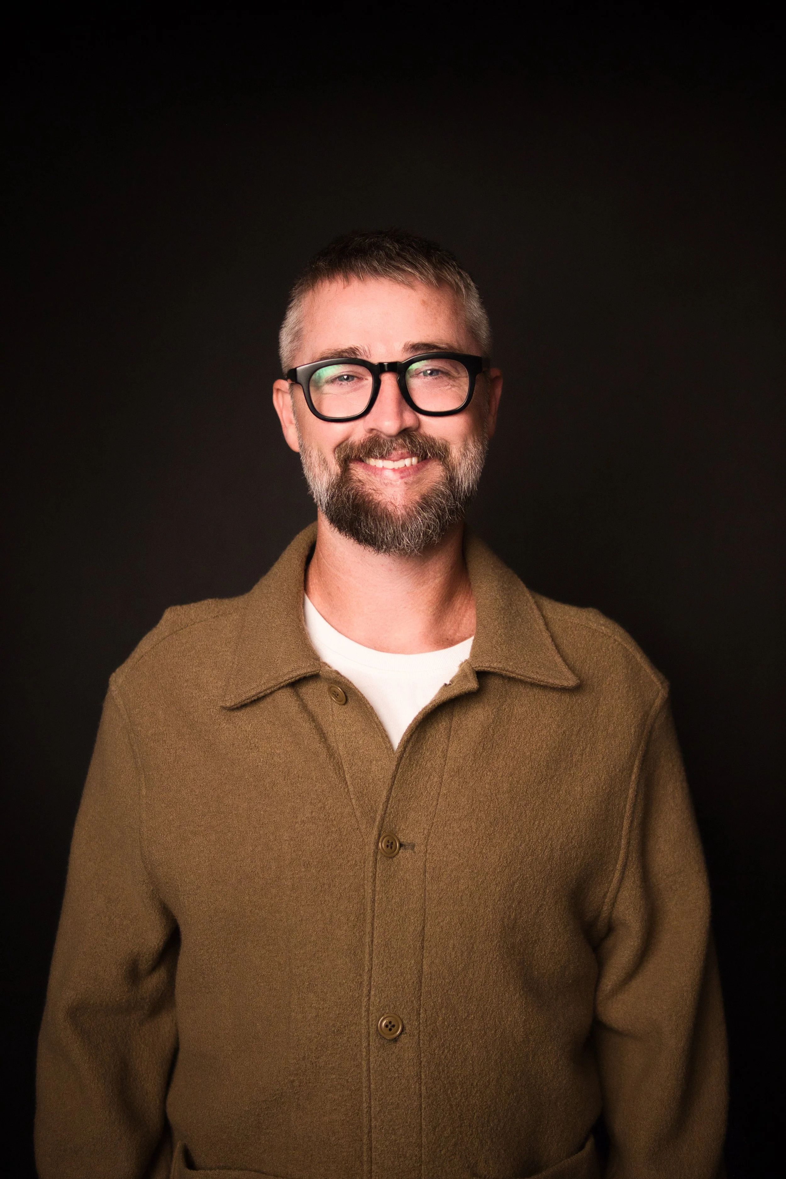 Portrait of a smiling man with glasses, a beard, wearing a brown coat over a white shirt, against a black background.