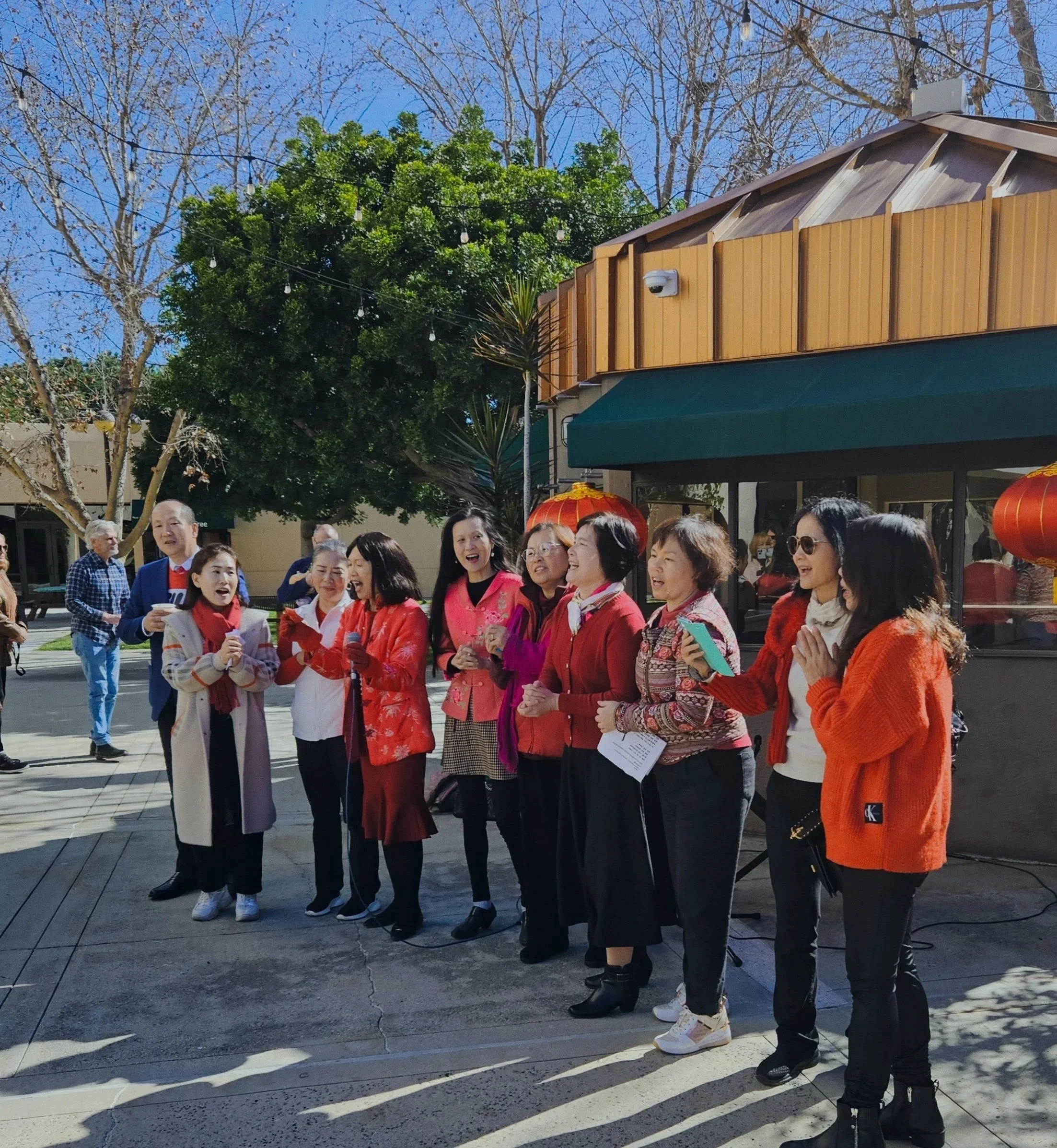 A group of diverse women standing outdoors on a sunny day, with some wearing traditional Asian clothing, chatting and smiling, with a background of trees, a building, and outdoor lanterns.