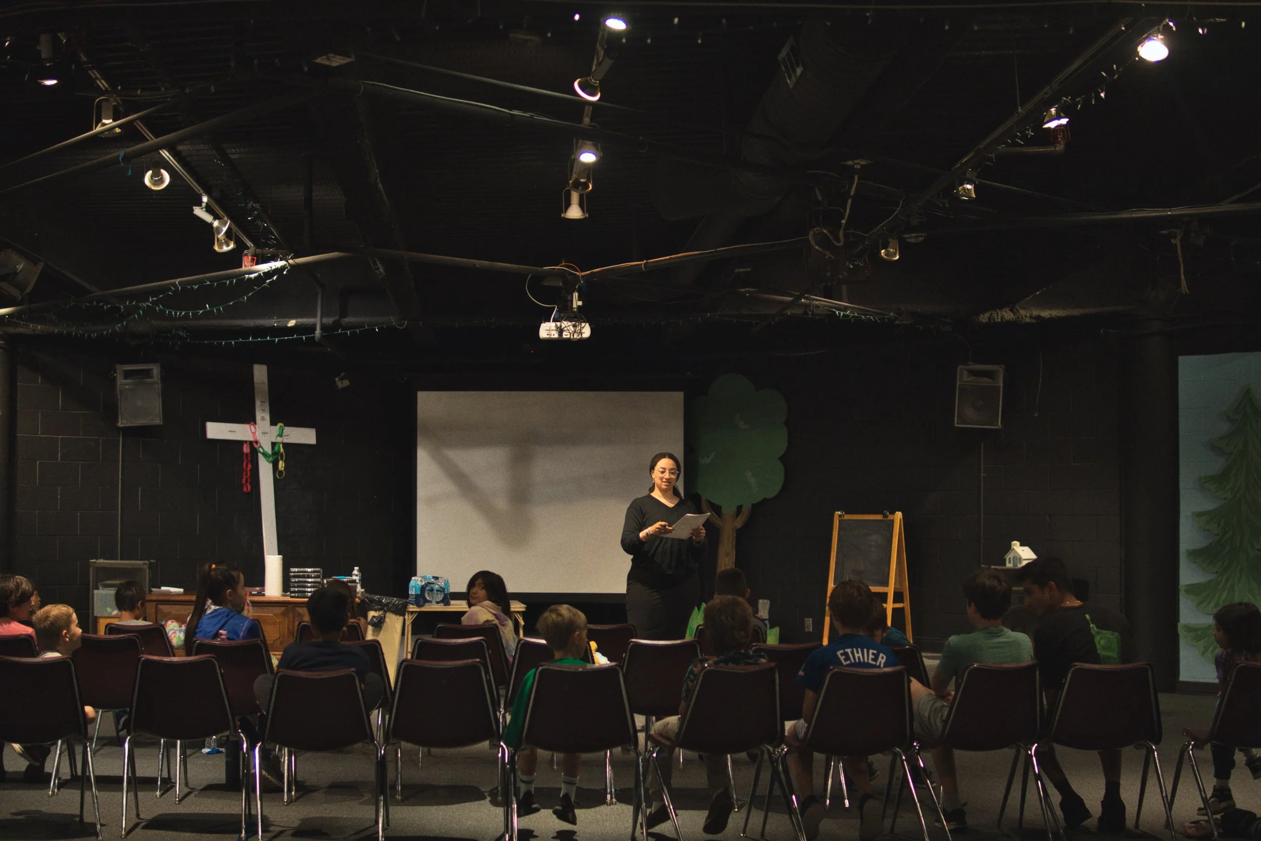 A woman standing in front of a large screen, holding papers, with children seated in chairs facing her inside a room with black walls and ceiling, decorated with lights and tree cutouts.