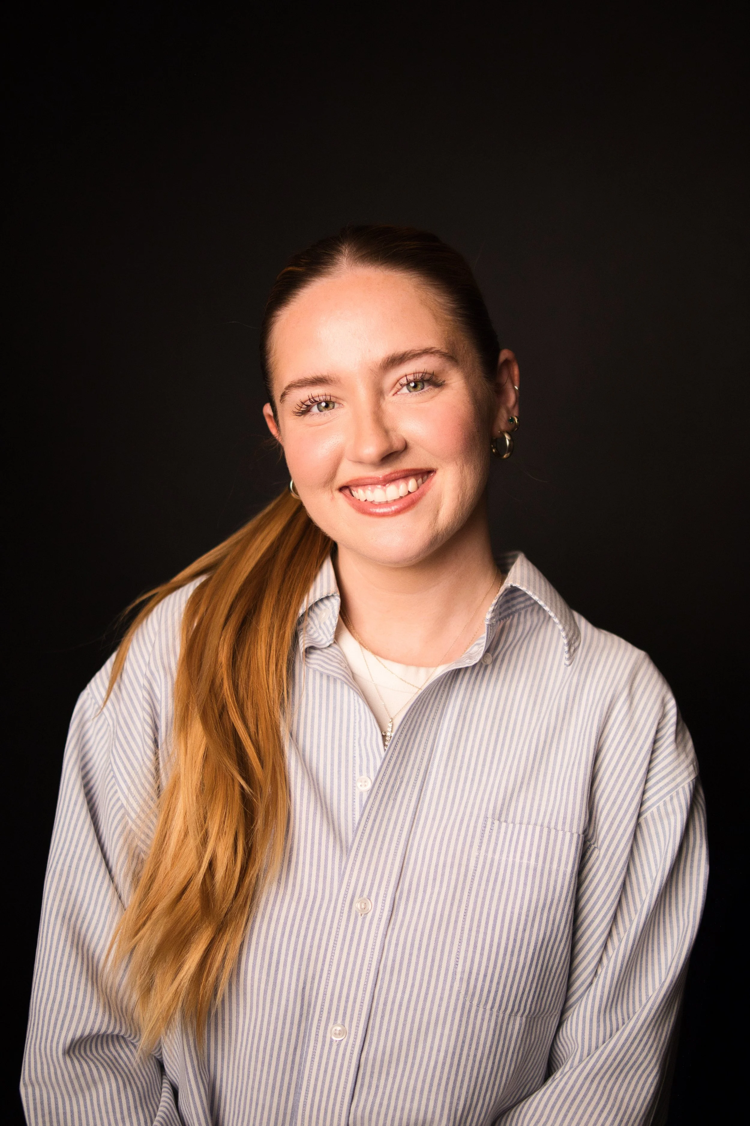 Close-up portrait of a young woman with long red hair, smiling, wearing a light striped button-up shirt, against a black background.