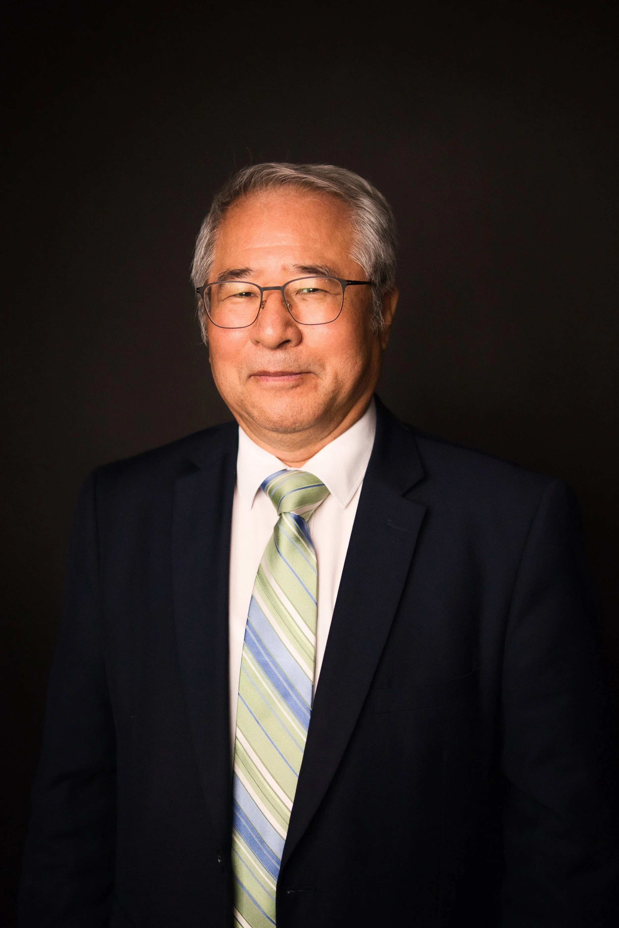 Portrait of an older Asian man wearing glasses, a dark suit, white shirt, and a light green striped tie, smiling gently against a dark background.