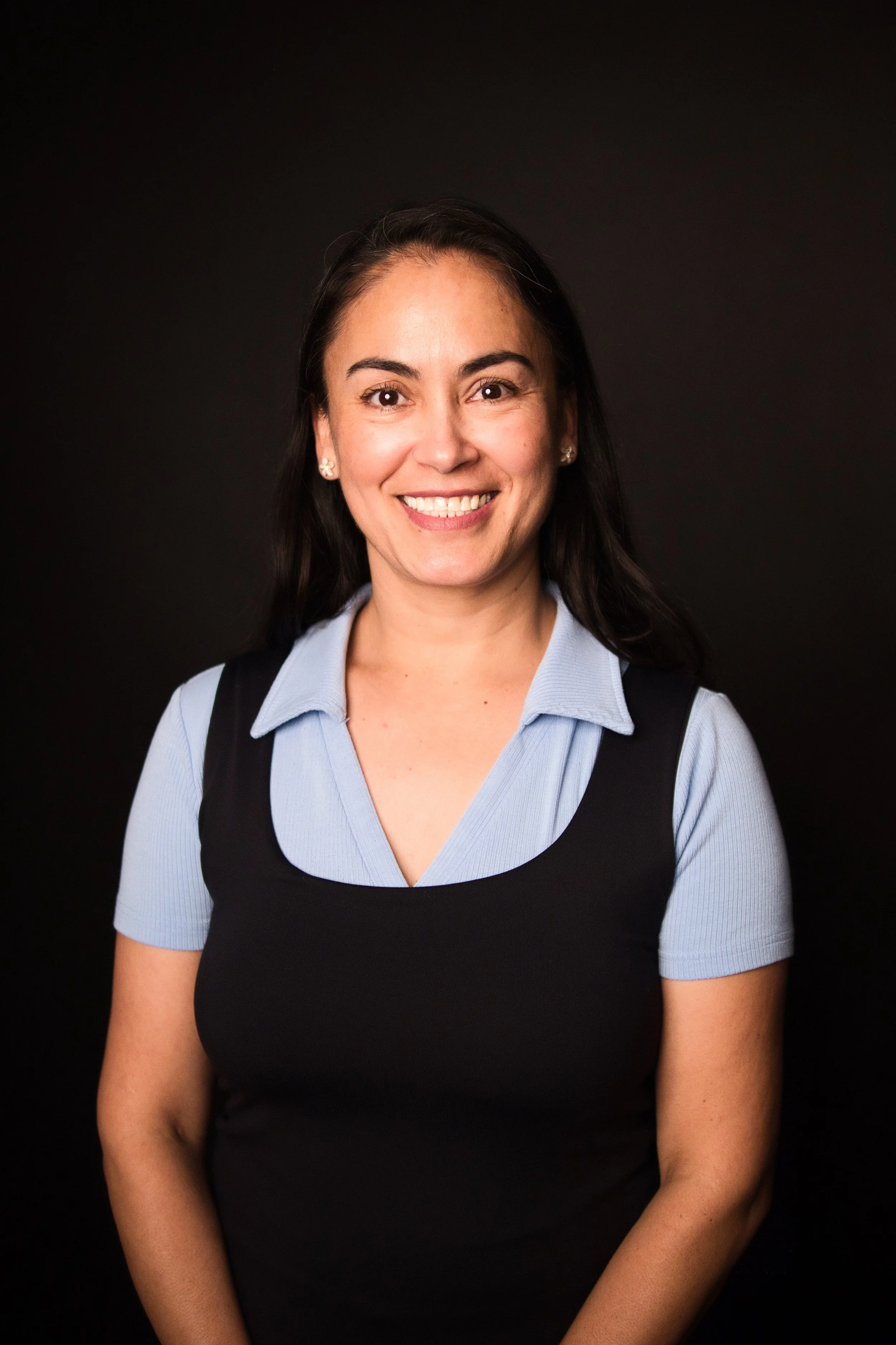 A woman with dark hair in a ponytail, wearing a light blue collared shirt under a black sleeveless top, smiling against a black background.