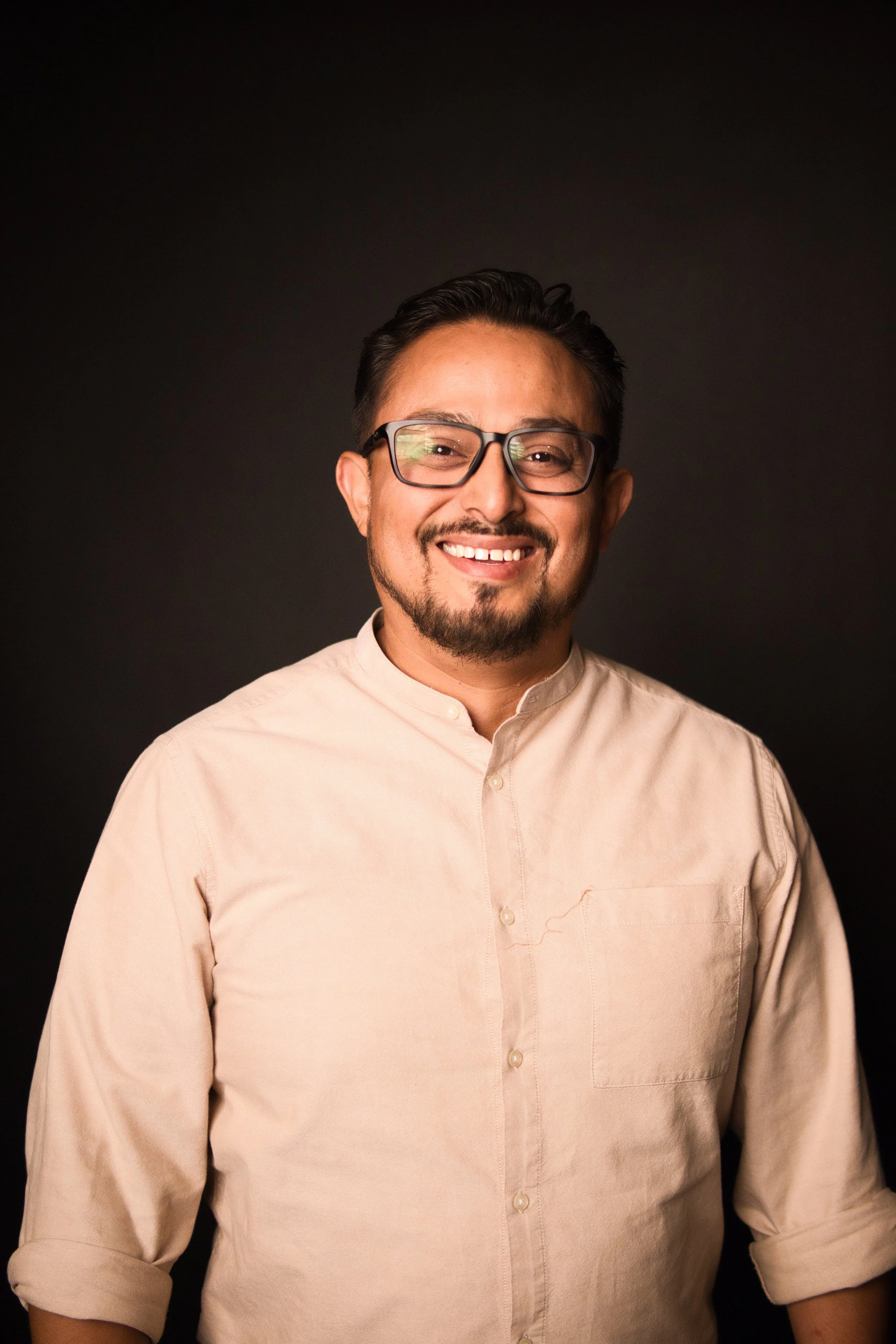 A smiling man with glasses, a dark hair styled back, a mustache and a beard in a light pink shirt against a black background.