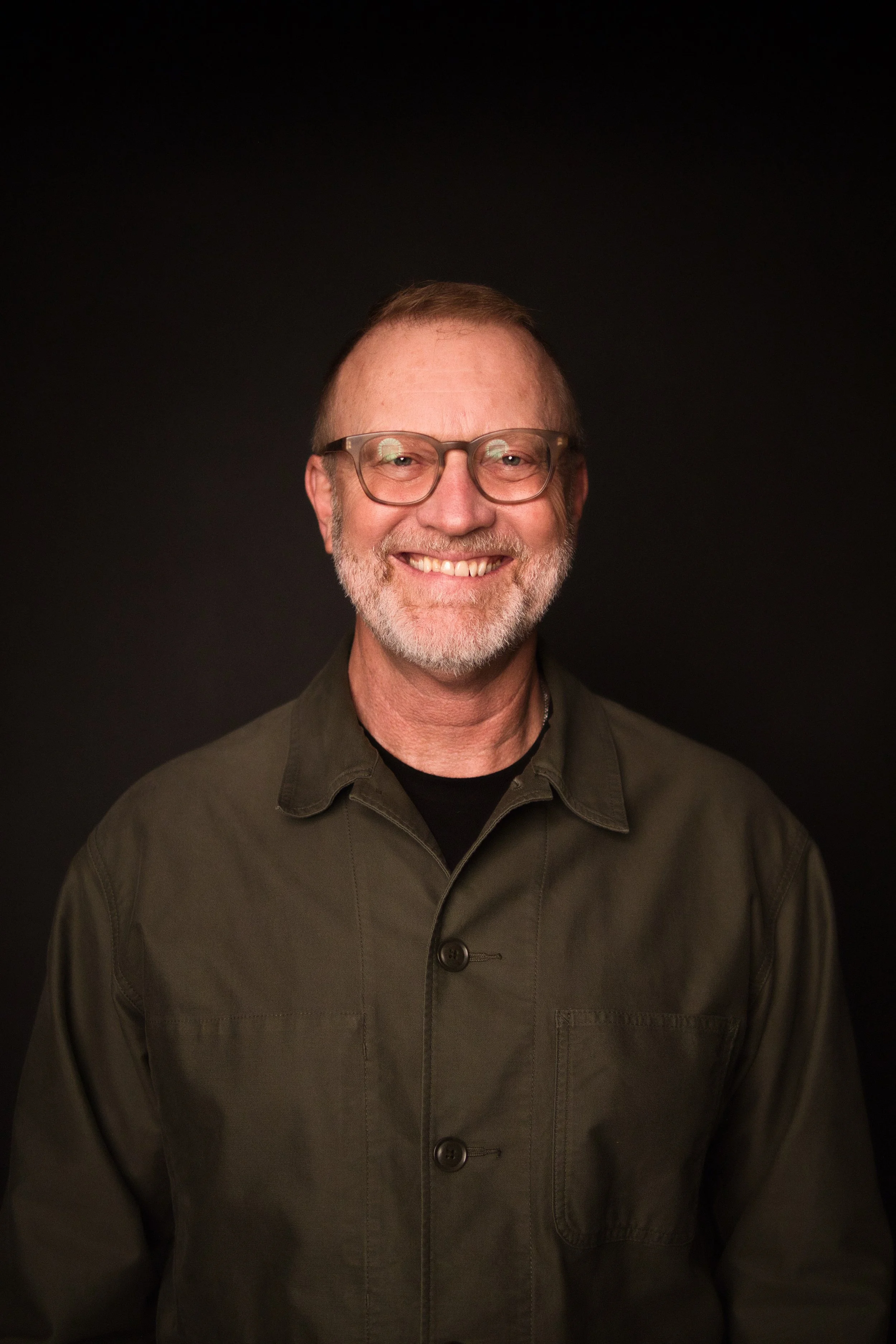 A middle-aged man with short, light brown hair, glasses, and a beard, smiling in front of a black background. He is wearing a dark green shirt.