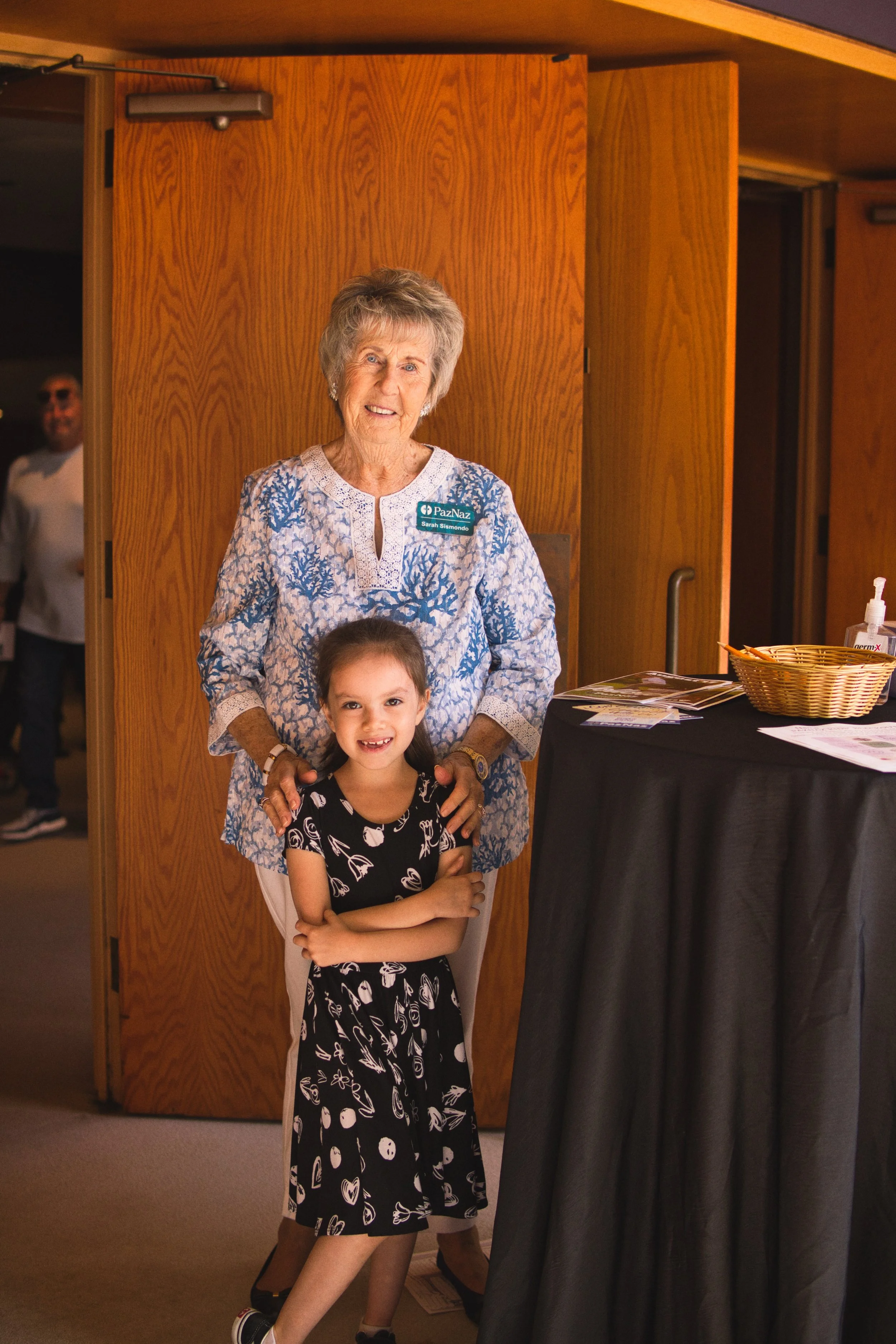 An elderly woman and a young girl standing in front of a wooden wall, smiling for the camera. The woman has short gray hair, is wearing a blue and white patterned blouse with a name tag, and has her arm around the girl. The girl has long dark hair, i