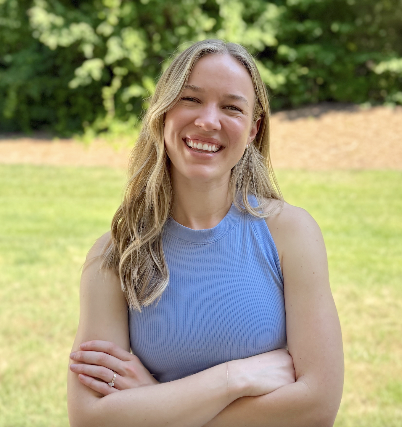 A woman with blonde hair wearing a blue sleeveless top, smiling outdoors with green foliage and grass in the background.