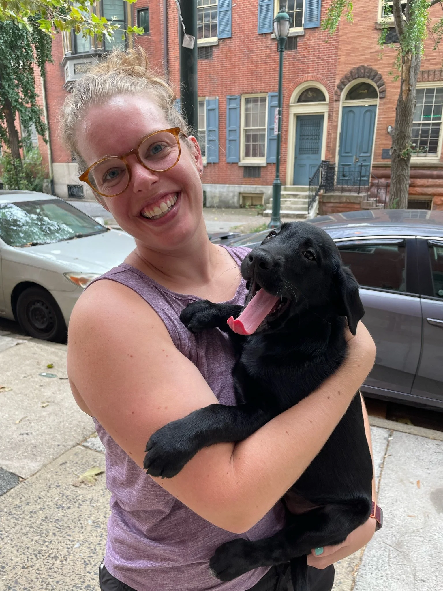 A smiling woman with light skin and glasses holding a black puppy outdoors on a city street with brick buildings, trees, parked cars, and a street lamp in the background.