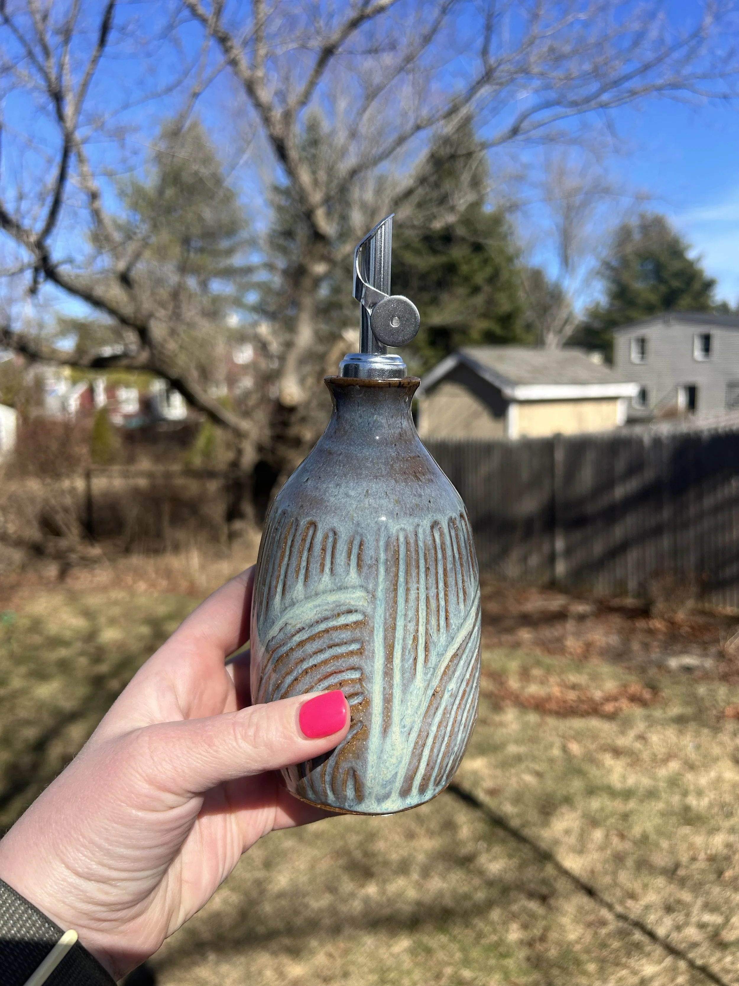 A hand with pink nail polish holding a ceramic lamp with a metal light fixture on top, outdoors with trees and houses in the background