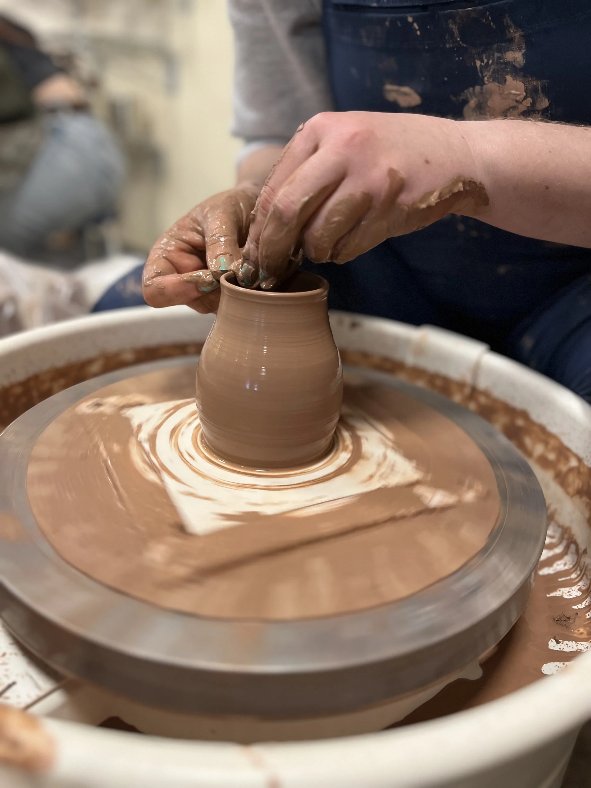 Person shaping a clay vase on a pottery wheel, hands covered in wet clay.