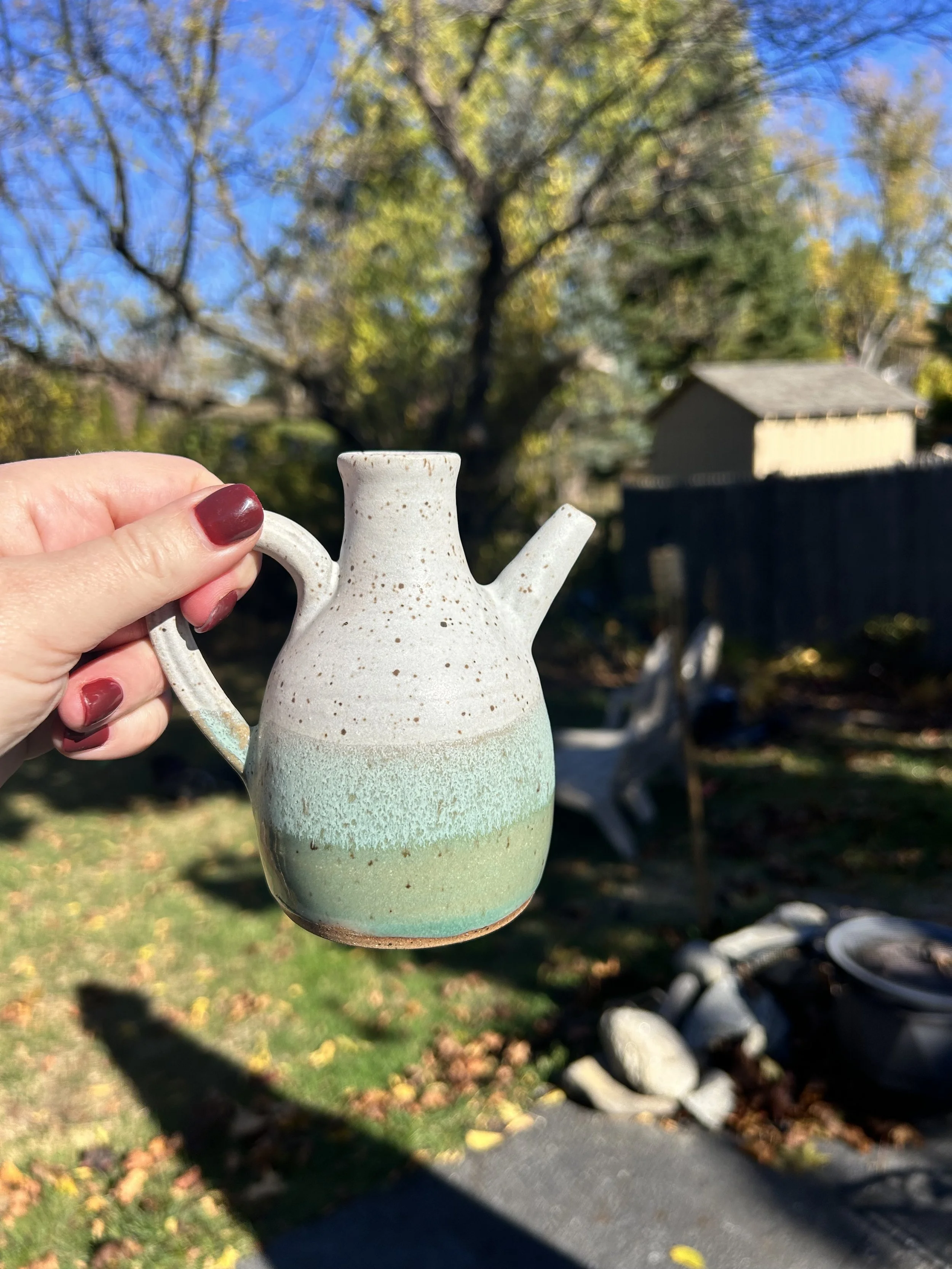 A person with red painted fingernails is holding a small, ceramic teapot with a speckled pattern, outdoors on a sunny day with trees and a house in the background.