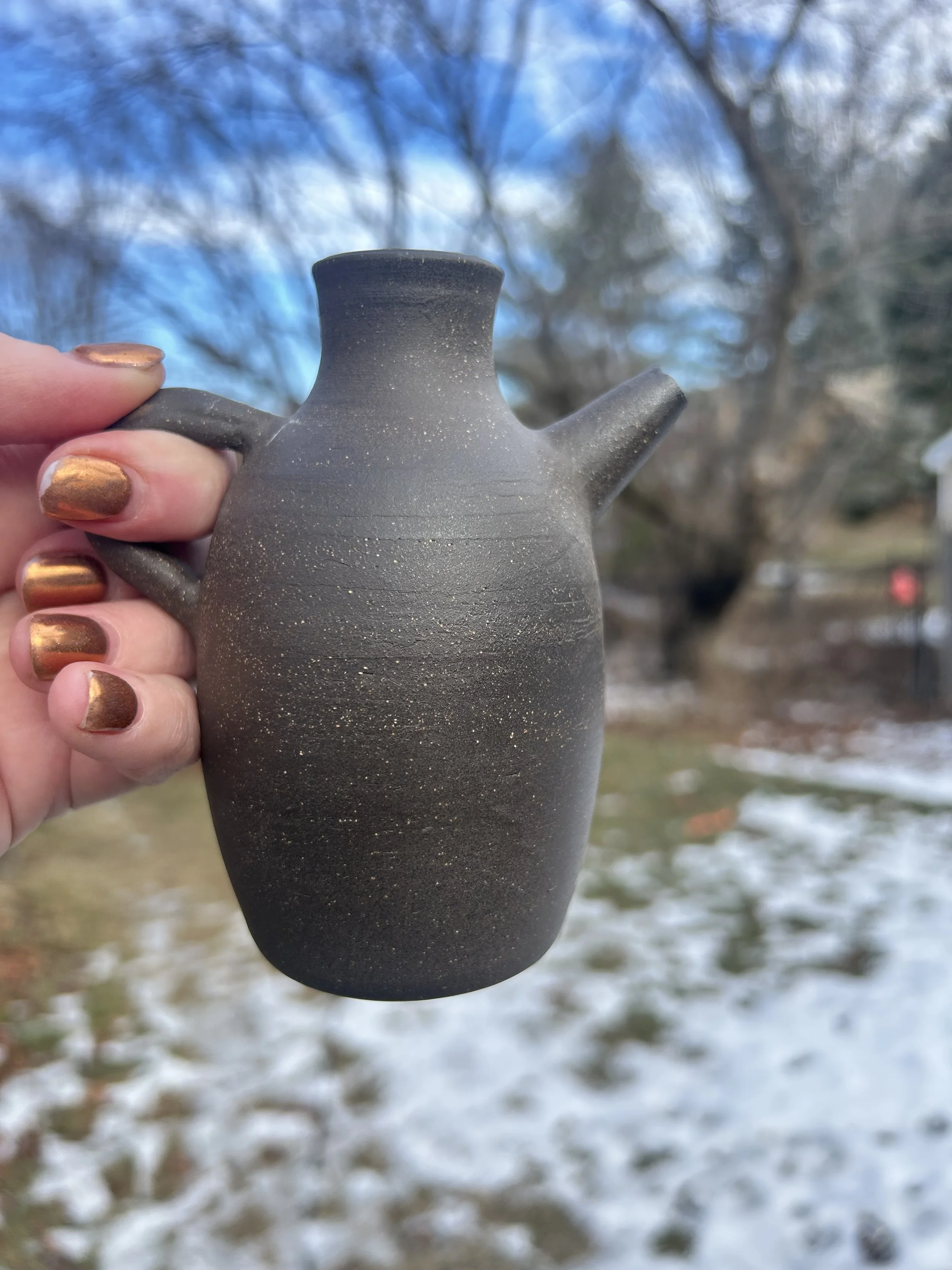 Hand holding a small, black, textured ceramic pitcher against a blurred outdoor background with trees and patches of snow on the ground.