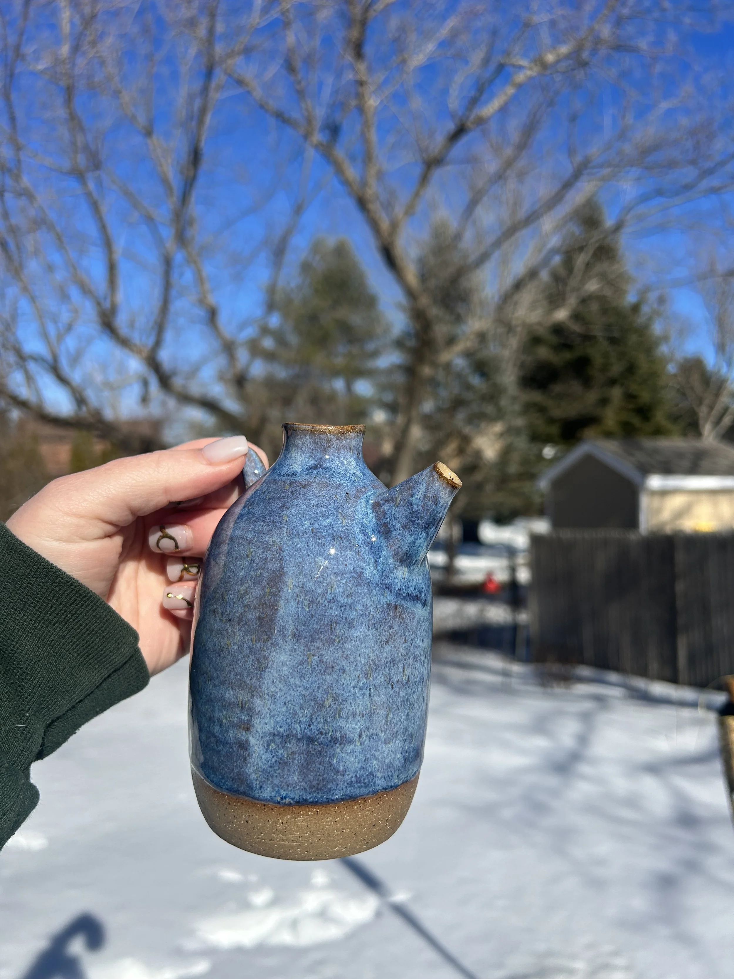 A person holding a blue and brown ceramic teapot outdoors during winter, with snow on the ground, leafless trees, and a building in the background.