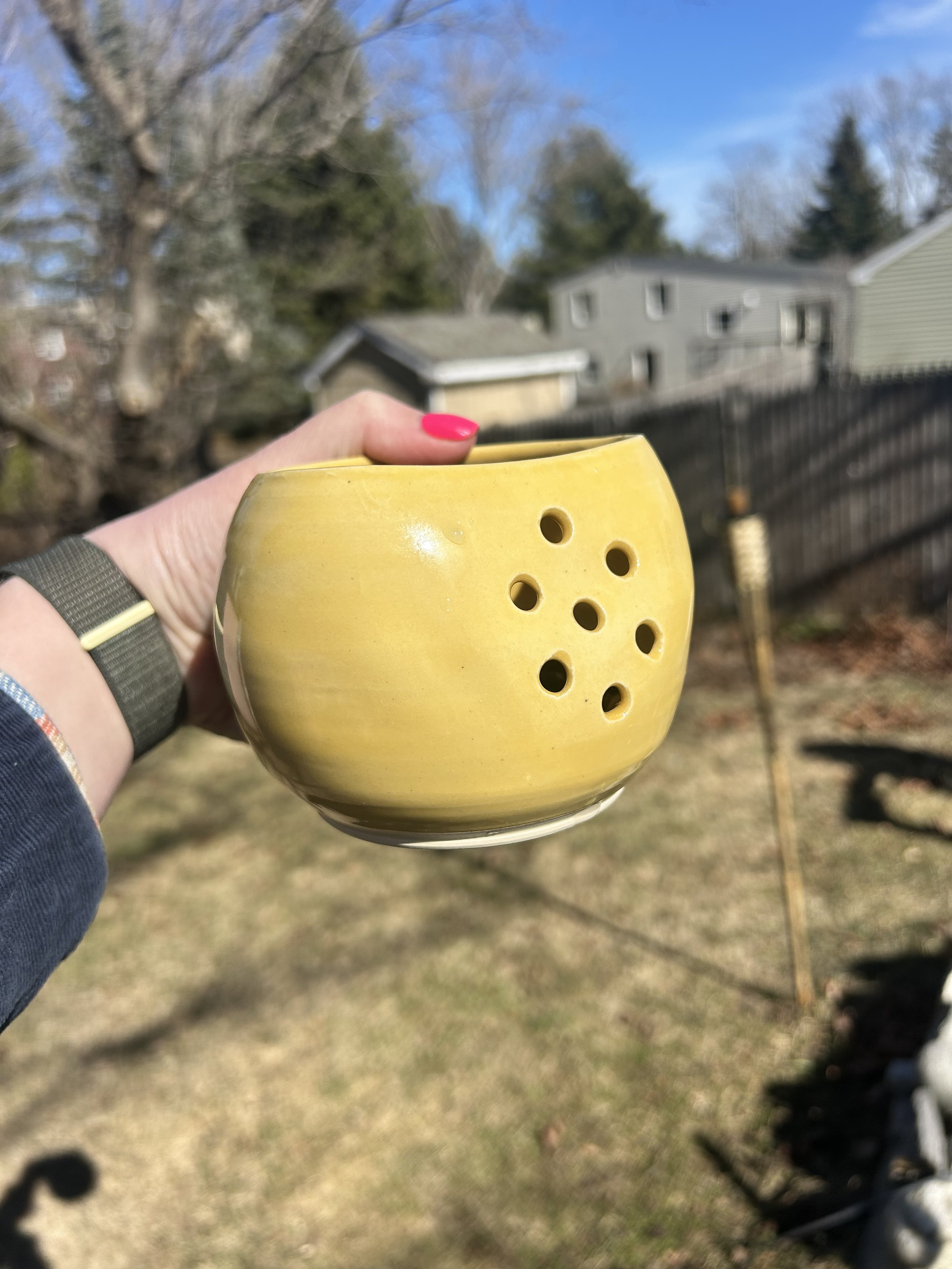 Close-up of a yellow ceramic hole puncher held in a person's hand outdoors, with a background of trees, houses, and a blue sky.