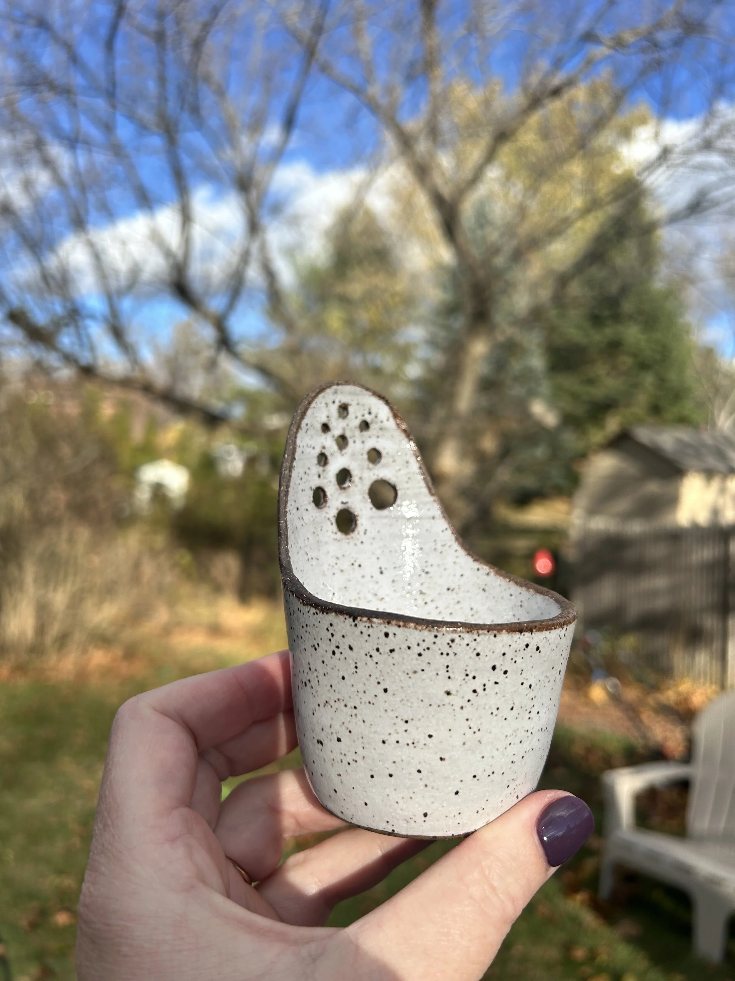 A hand holding a speckled ceramic watering can with a large spout, with a background of trees and a partly cloudy sky.