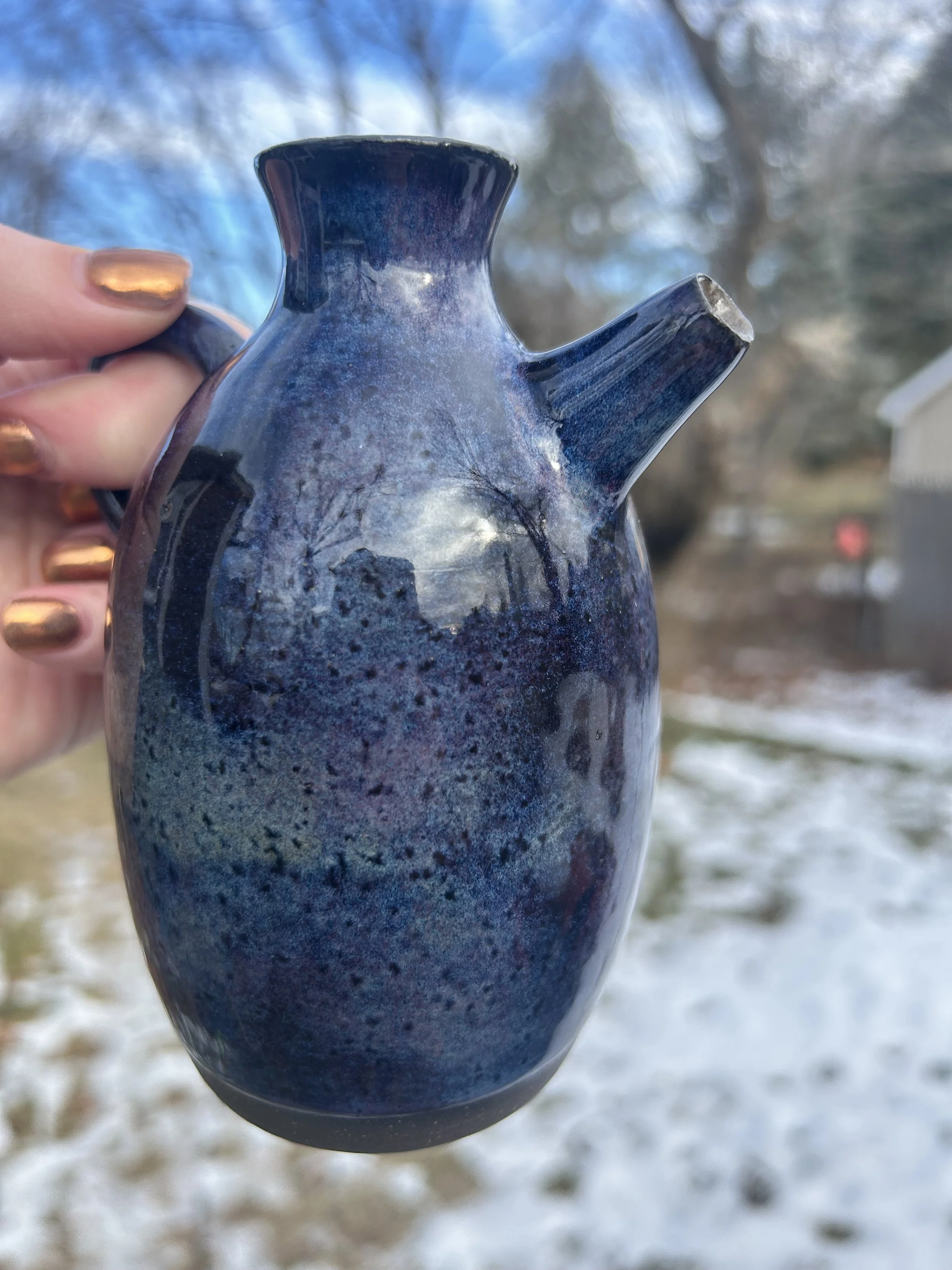 Person holding a blue ceramic teapot outdoors with trees and a house in the background.
