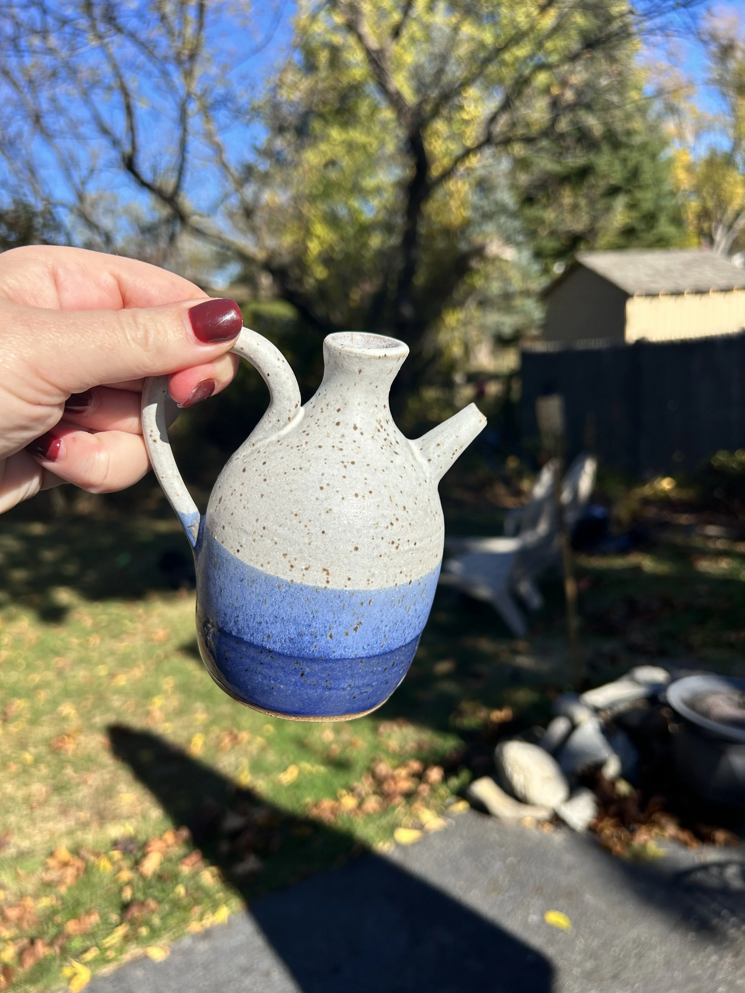 White ceramic watering can with a blue gradient at the bottom, being held by a hand with red nail polish, outdoors on a sunny day with trees and a building in the background.