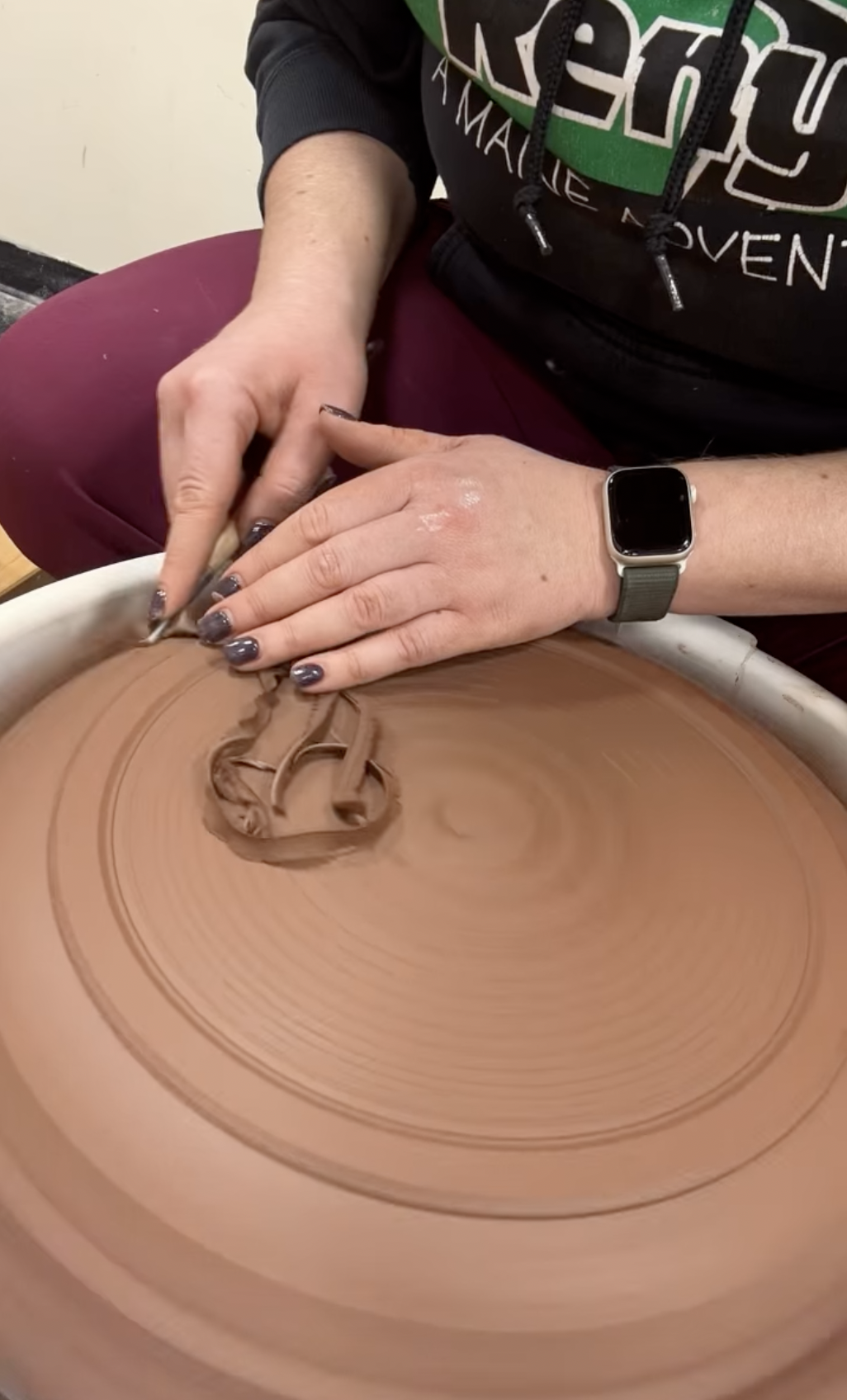 Person shaping clay on a pottery wheel, with hands covered in clay and wearing a smartwatch.