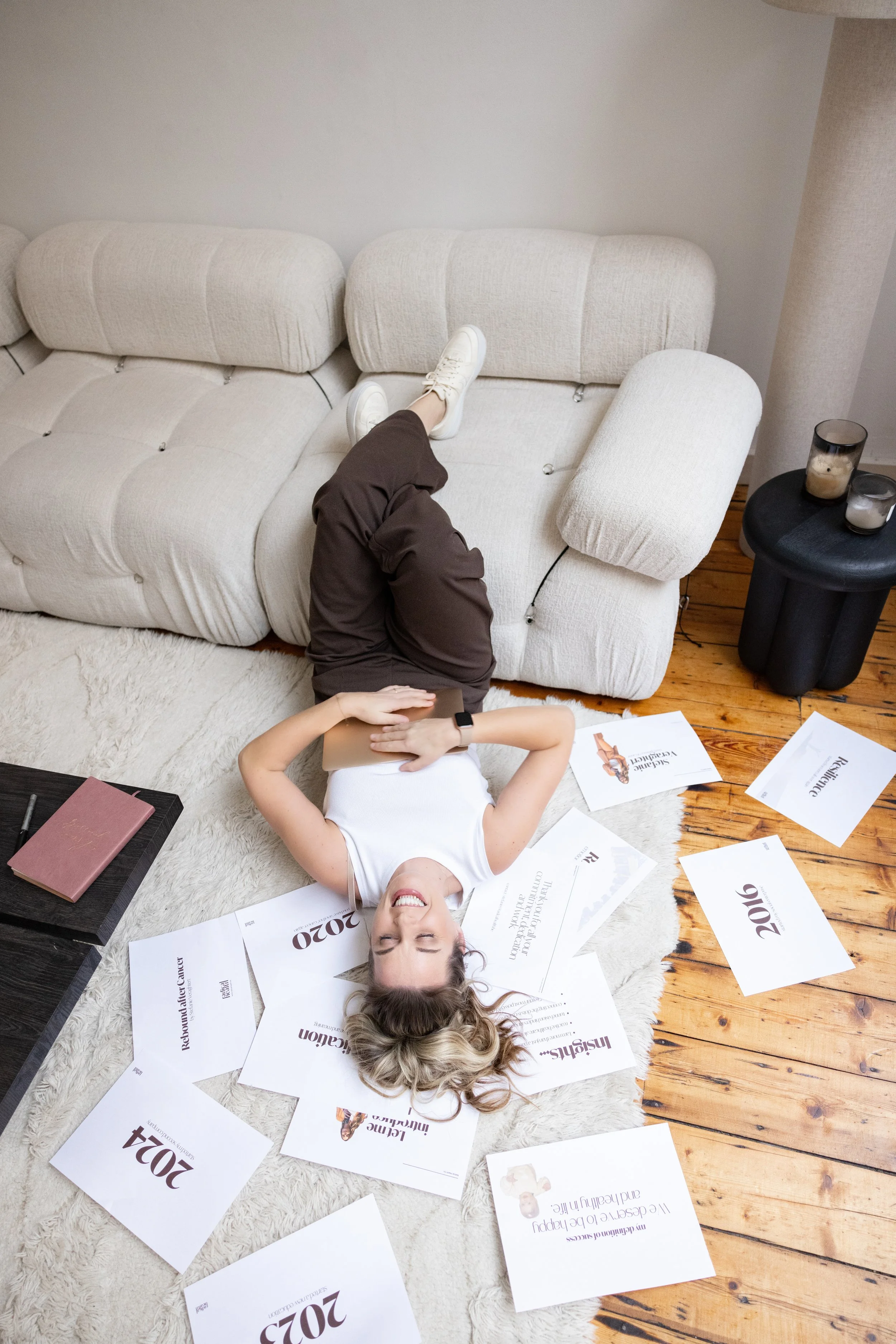 Vrouw ligt op de vloer met haar hoofd op de grond, omringd door papieren met jaartallen en tekst, terwijl ze op haar rug ligt met een glimlach.