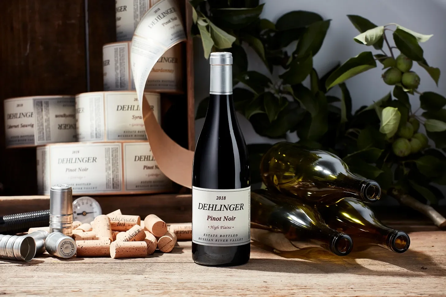 A bottle of Dehlinger 2018 Pinot Noir wine standing upright on a wooden surface, with three empty wine bottles on their sides behind it, wine corks and wine corking tools in front, a box of Dehlinger wine in the background, and a leafy green plant.