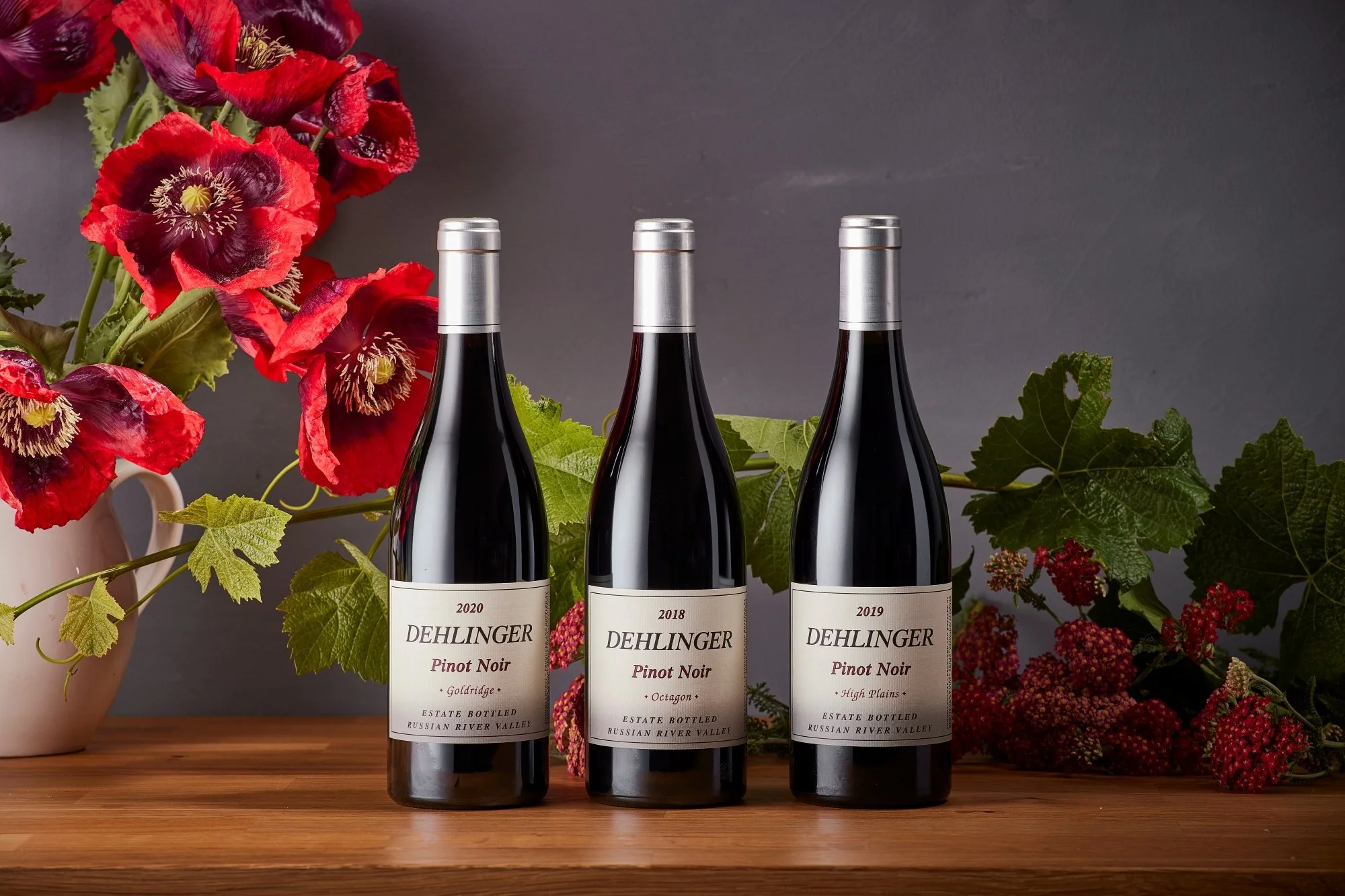 Three bottles of Dehlinger Pinot Noir wine with vintage years 2020, 2018, and 2019, set on a wooden surface surrounded by red flowers and green leaves against a gray background.