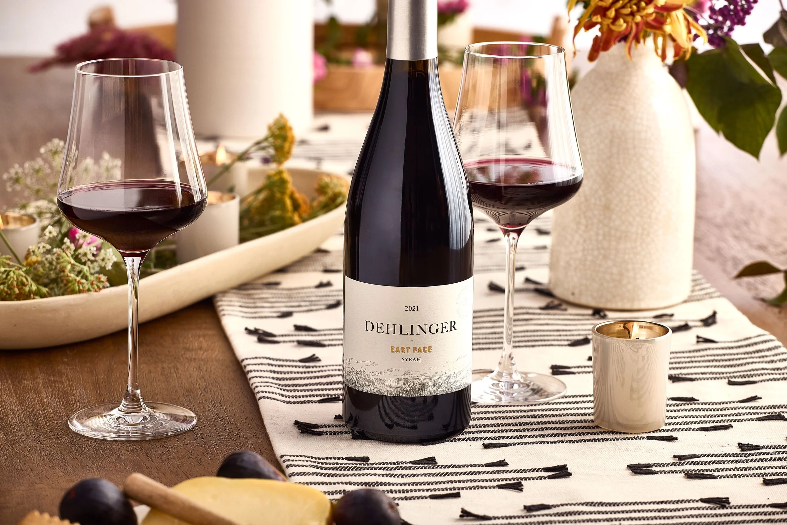 A bottle of Dehlinger East Face Syrah wine with two glasses of red wine, a white candle, and a vase with flowers on a dining table.