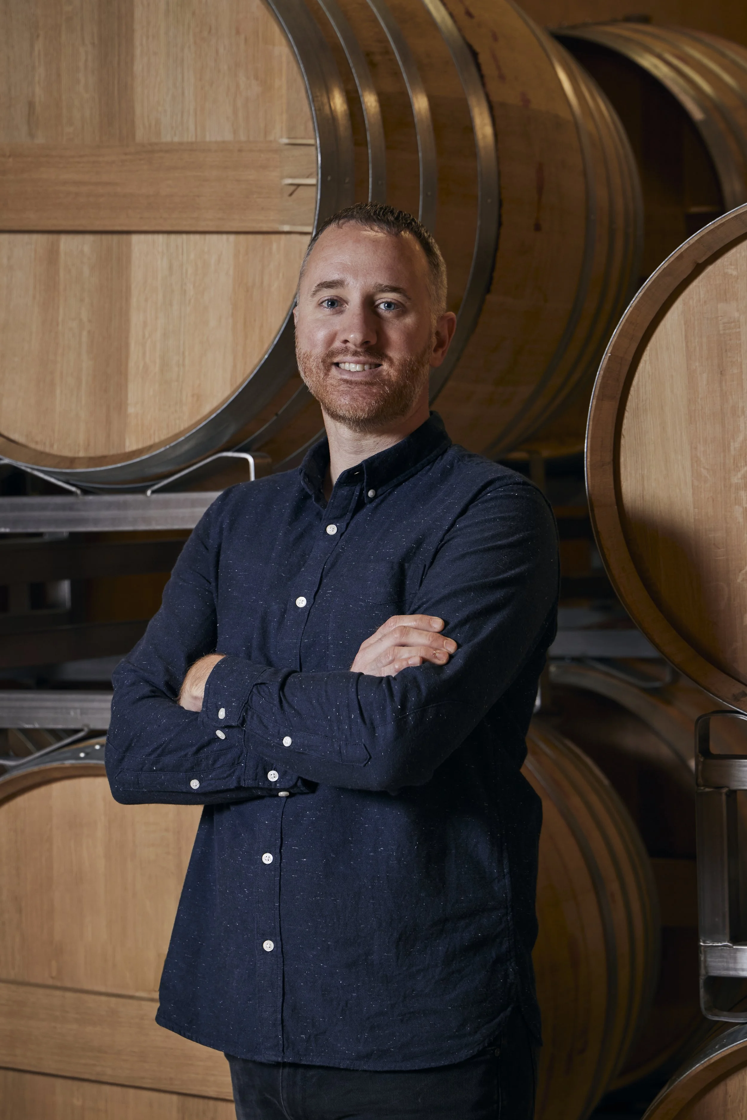 A man with short hair and a beard wearing a dark blue button-up shirt, standing with arms crossed in front of a backdrop of wine barrels in a winery or cellar.