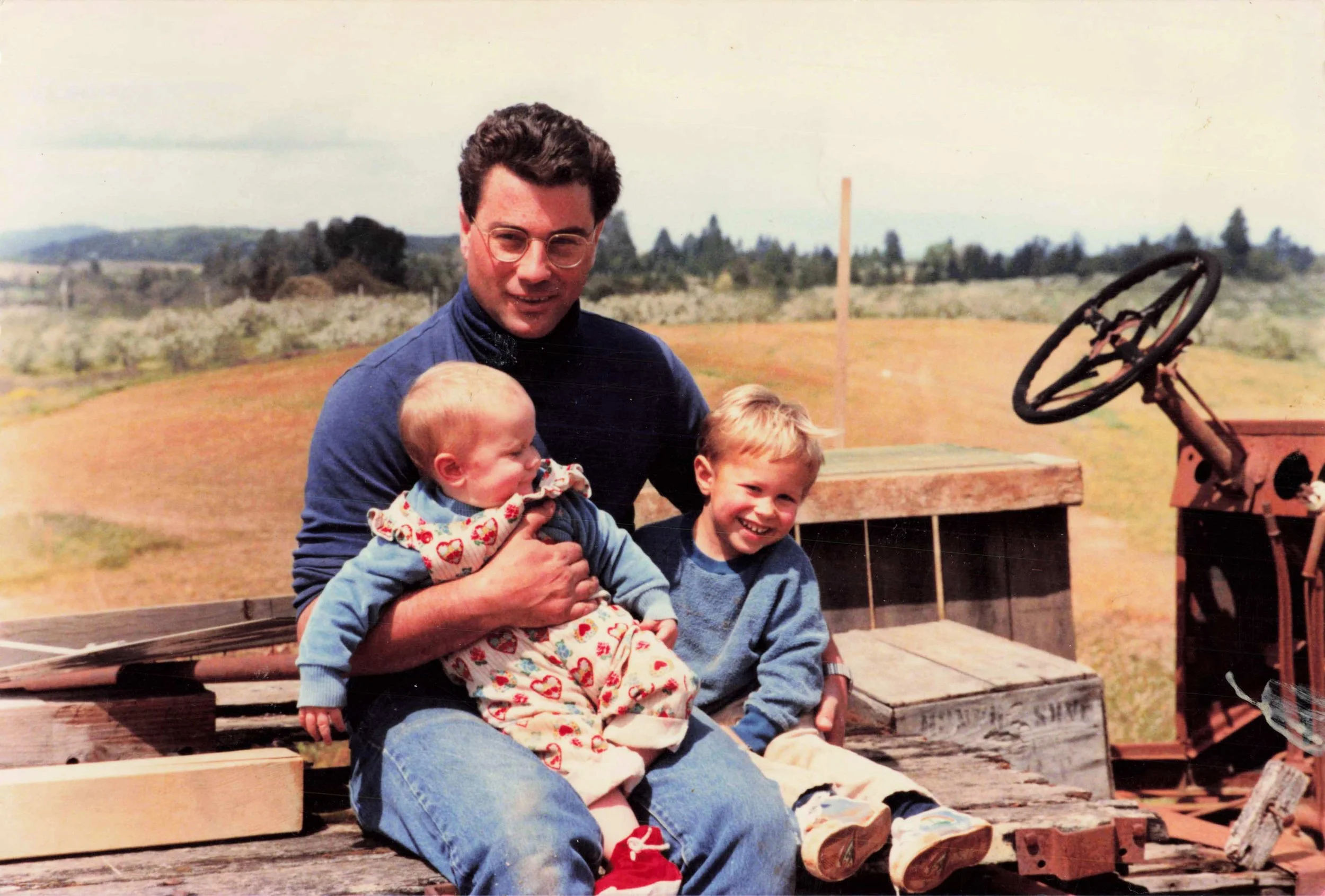 A man with glasses holding a young girl and a boy, sitting on wooden train cars outdoors with a rural landscape in the background.