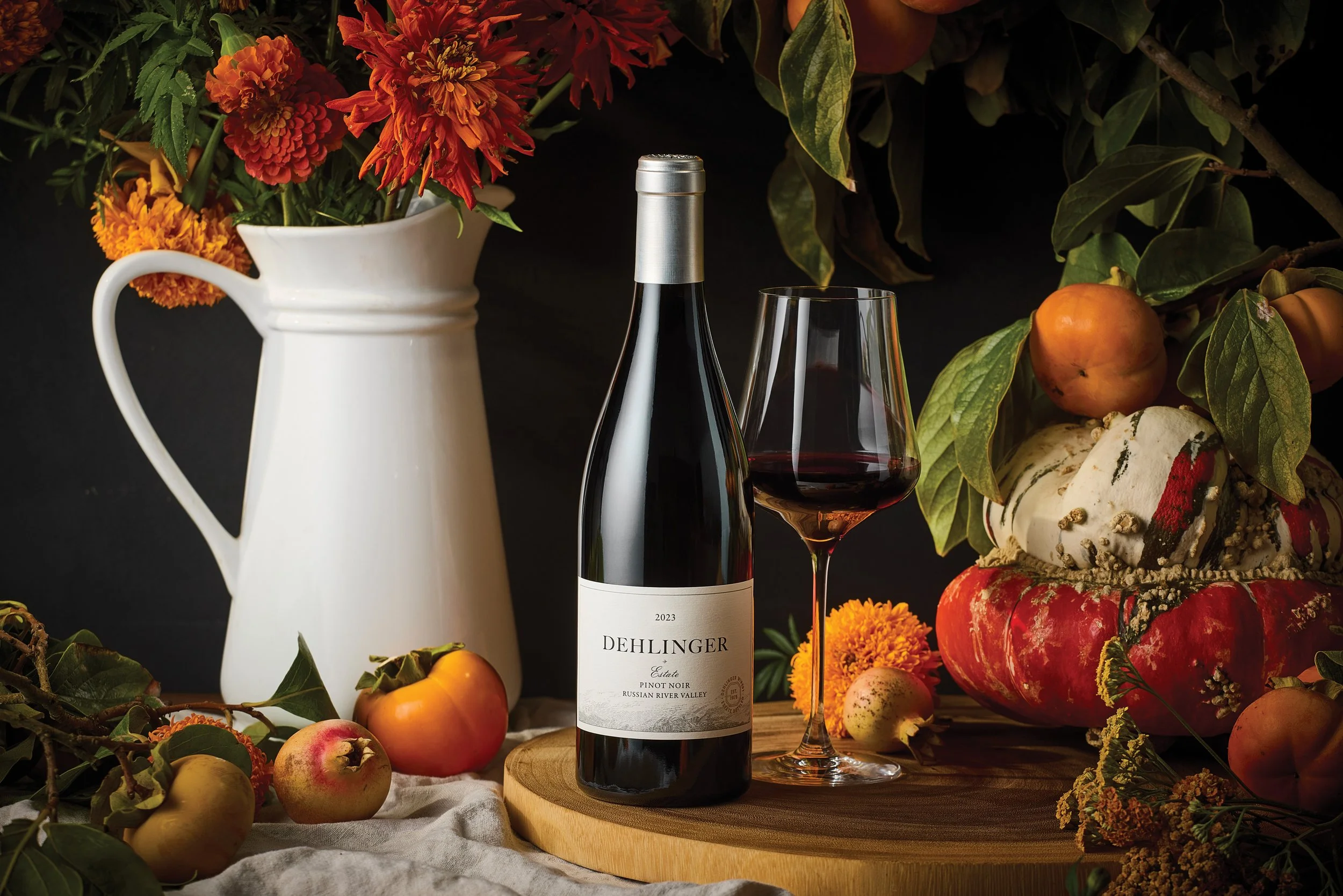 A bottle of red wine labeled 'Dehlinger Estate Pinot Noir Russian River Valley' with a glass of red wine beside it, surrounded by a white pitcher of bright orange and red flowers, various apples, and pumpkins on a wooden and cloth surface, with green leaves and branches in the background.