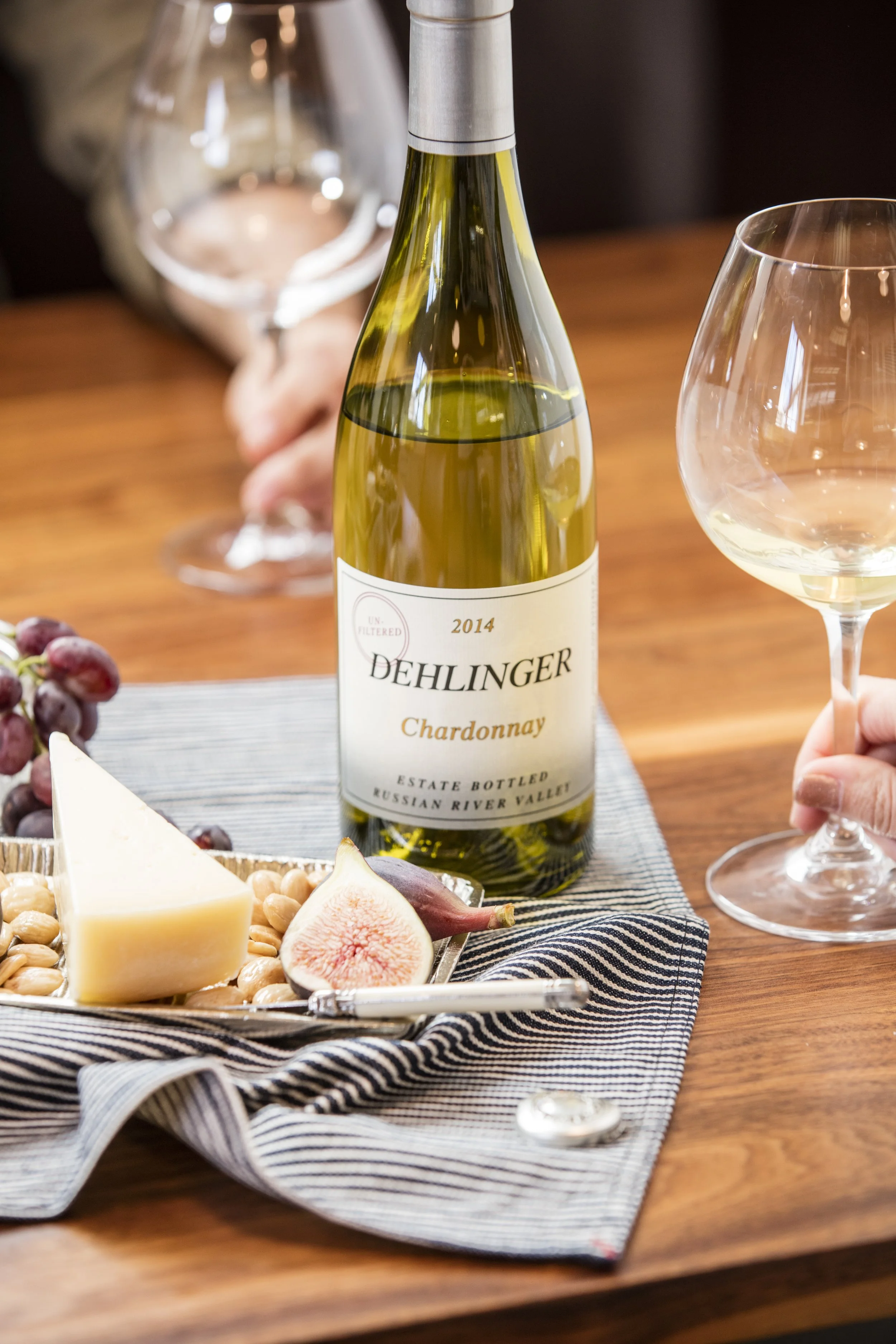 A table setting with a bottle of Dehleringer Chardonnay wine, two empty wine glasses, a cheese plate with cheese, grapes, and figs on a striped cloth.