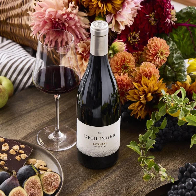 A bottle of Dehlinger Pinot Noir wine, a glass of red wine, and a plate of assorted fruits including figs and grapes, surrounded by colorful flowers and green foliage on a wooden table.