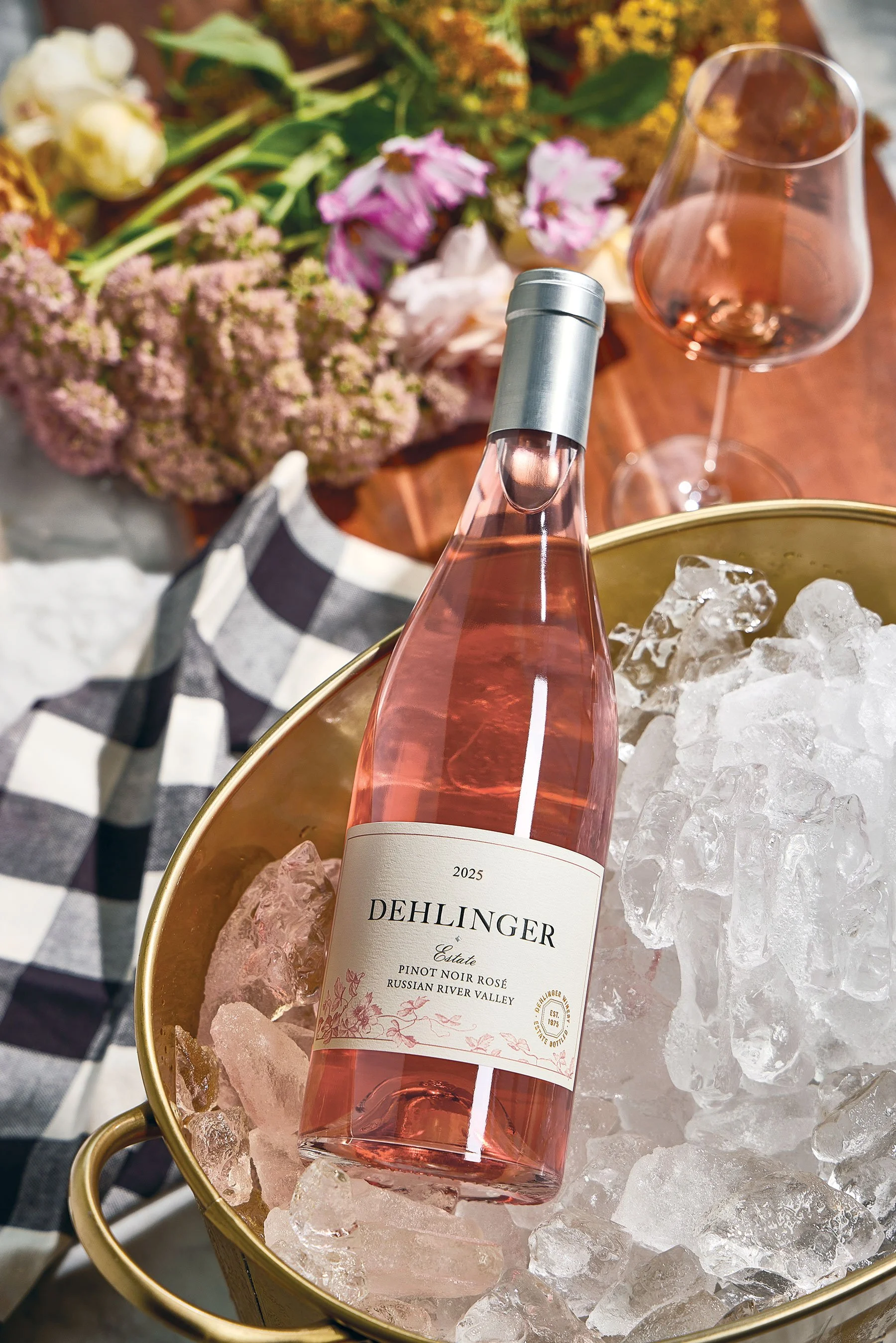 A bottle of Dehlinger Estate Pinot Noir Rosé wine on ice in a gold bucket, with a wine glass of rosé behind it, and a bouquet of flowers in the background.